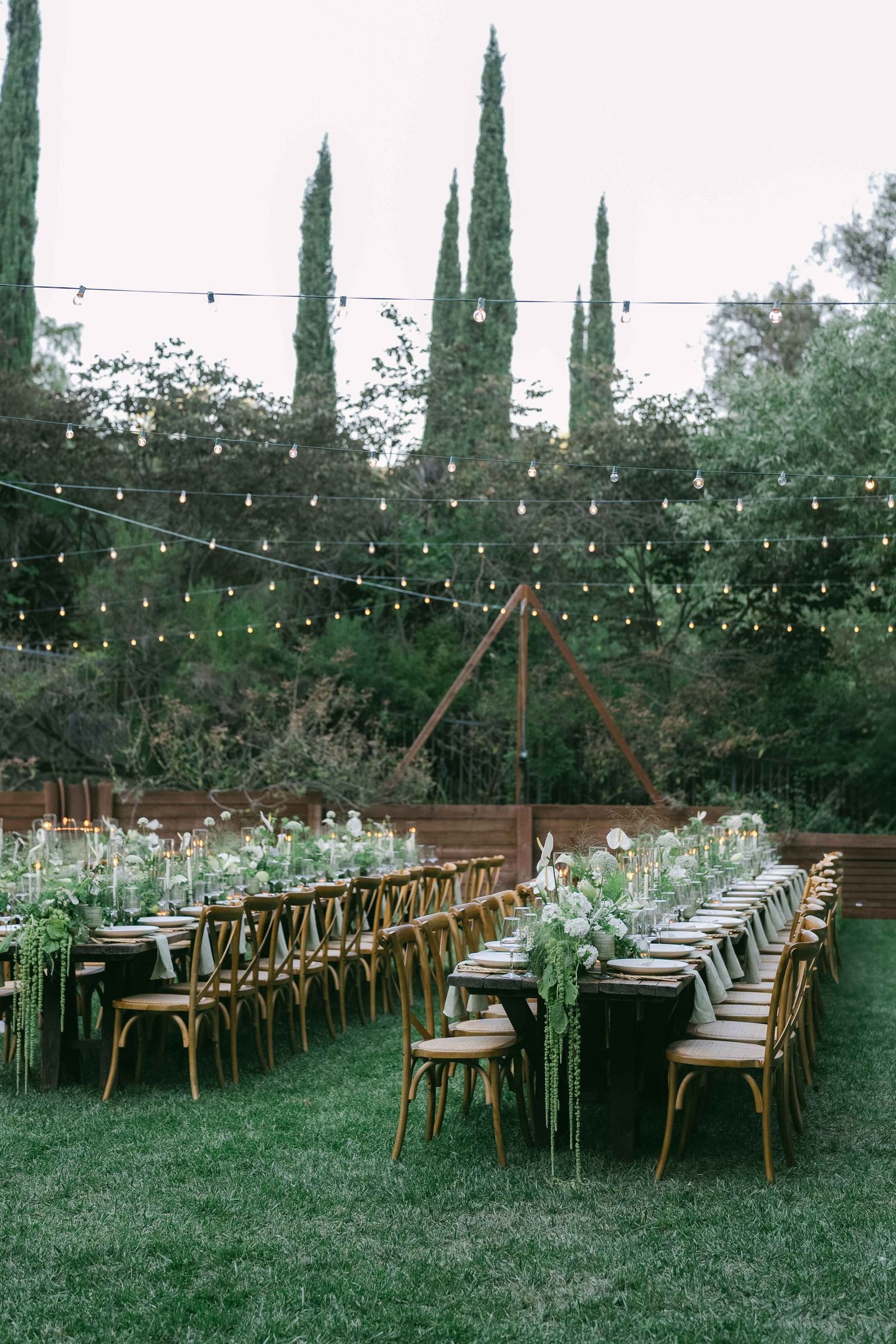 Garden wedding reception with long tables, cross-back chairs and overhead string lights framed by cypress trees.