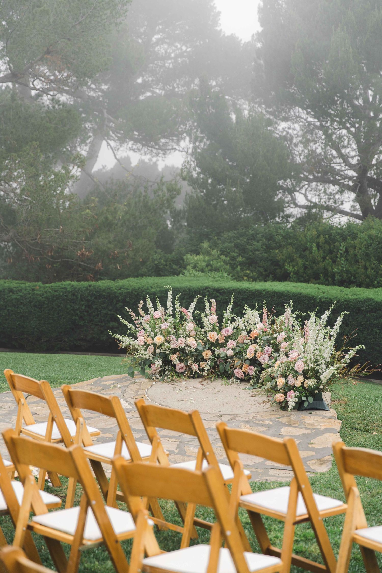 Outdoor wedding ceremony setup with wooden chairs and floral arrangements in misty garden setting.