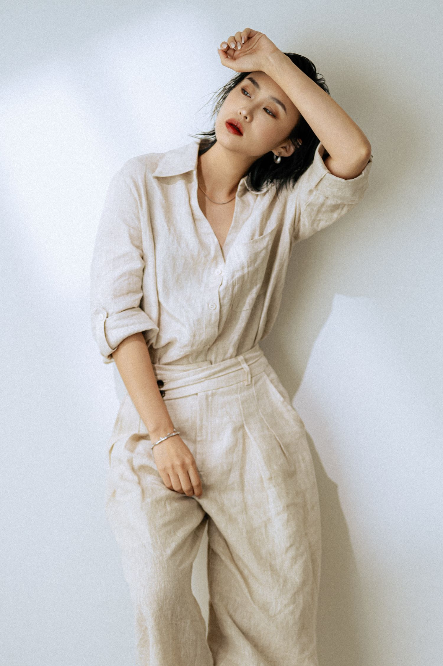 Artistic fashion pose in all-cream outfit with dramatic lighting against white wall.