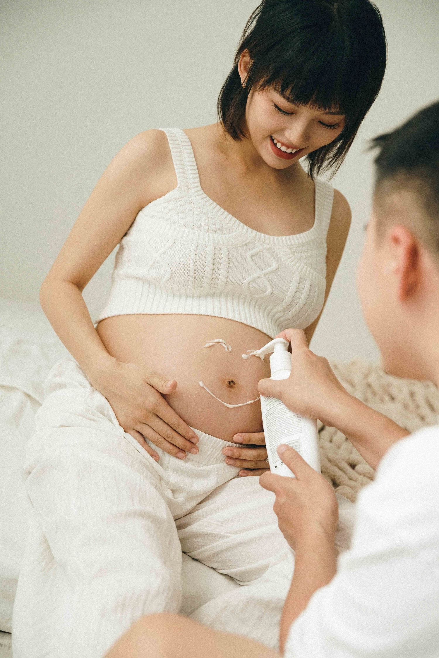 Person in white top showing pregnancy belly while sitting on bed.