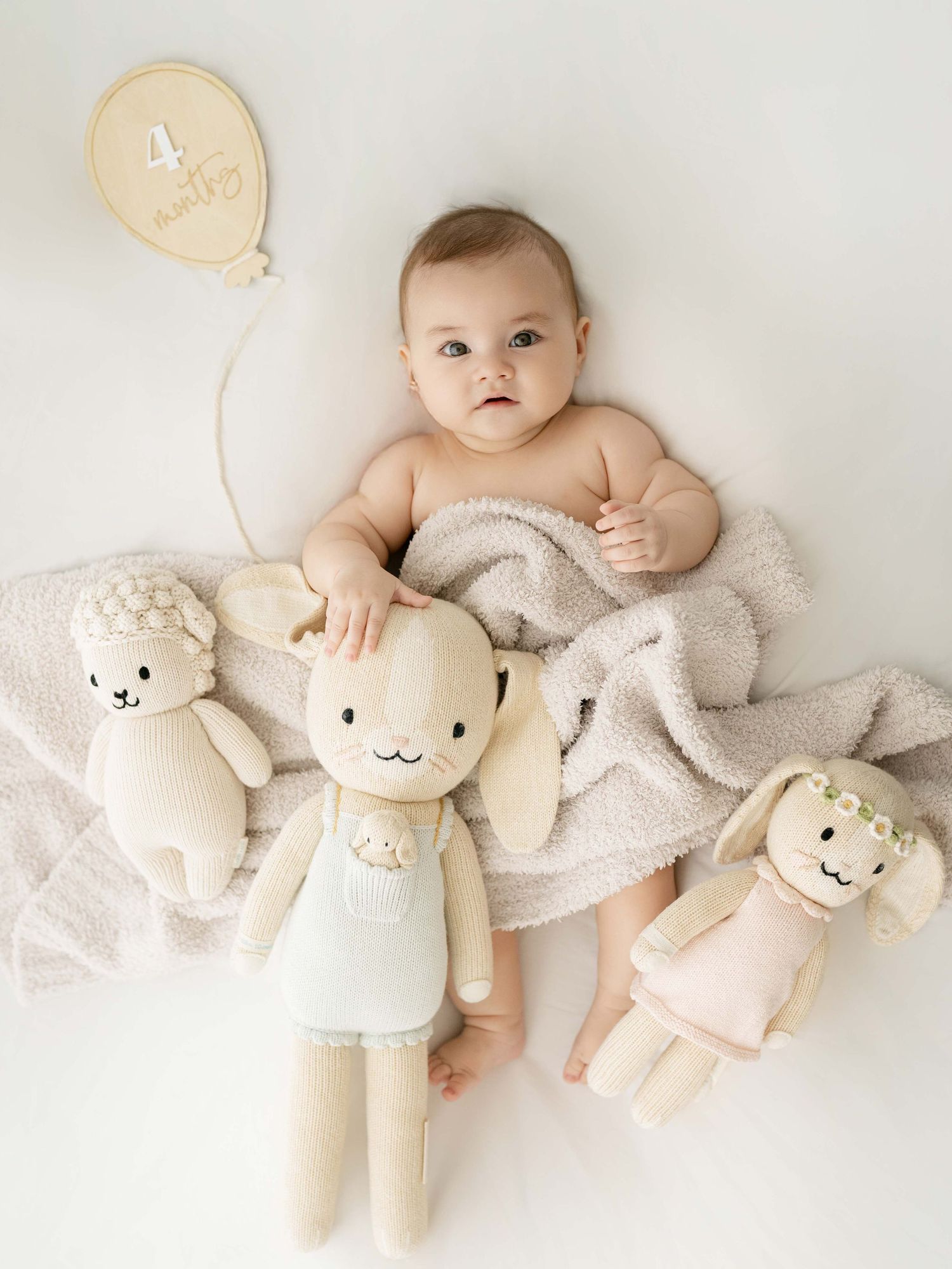 Baby resting on white blanket with plush bunny toys and milestone marker.
