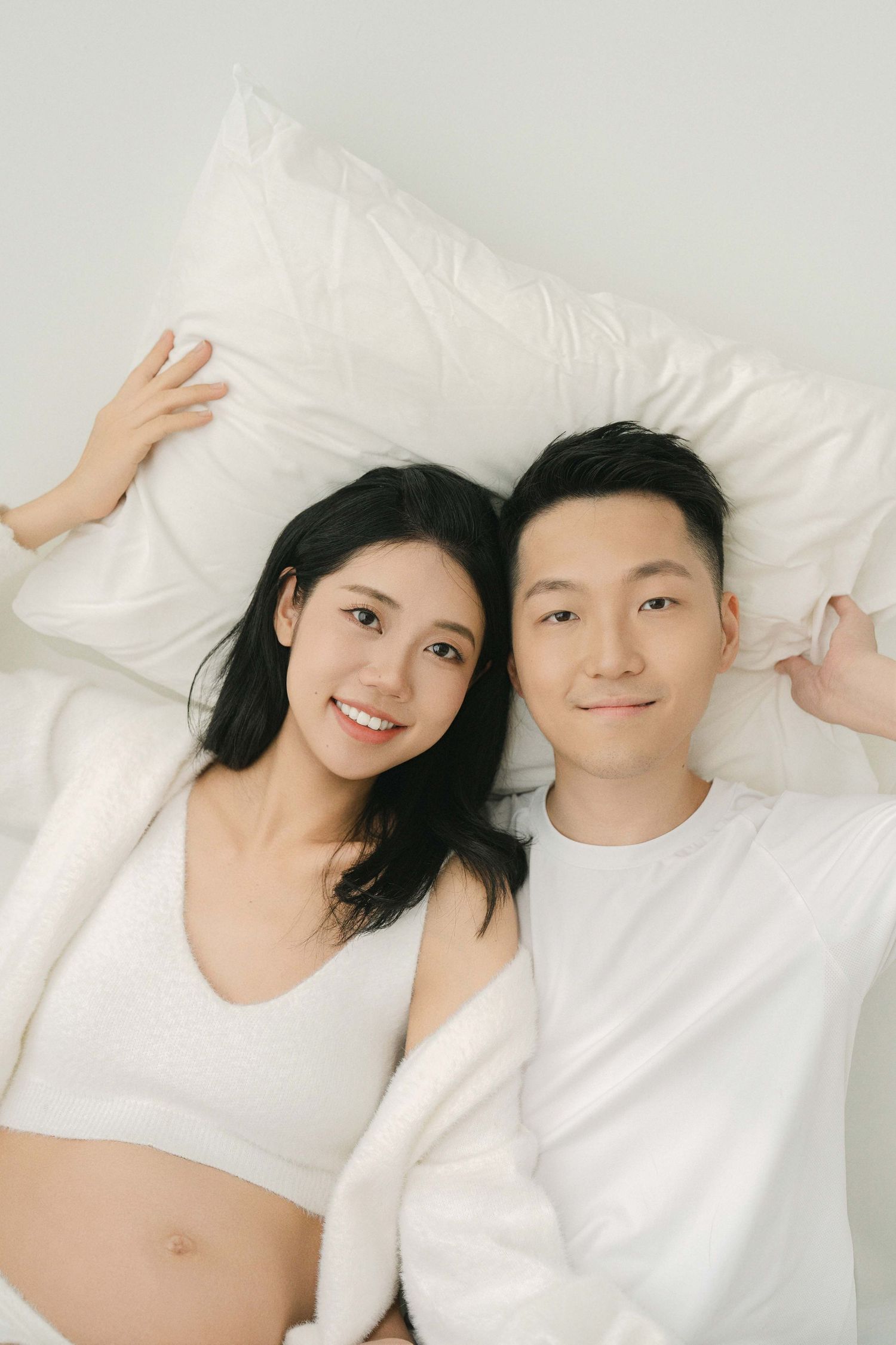 A couple in white clothing lying together on white bedding in a minimalist setting.