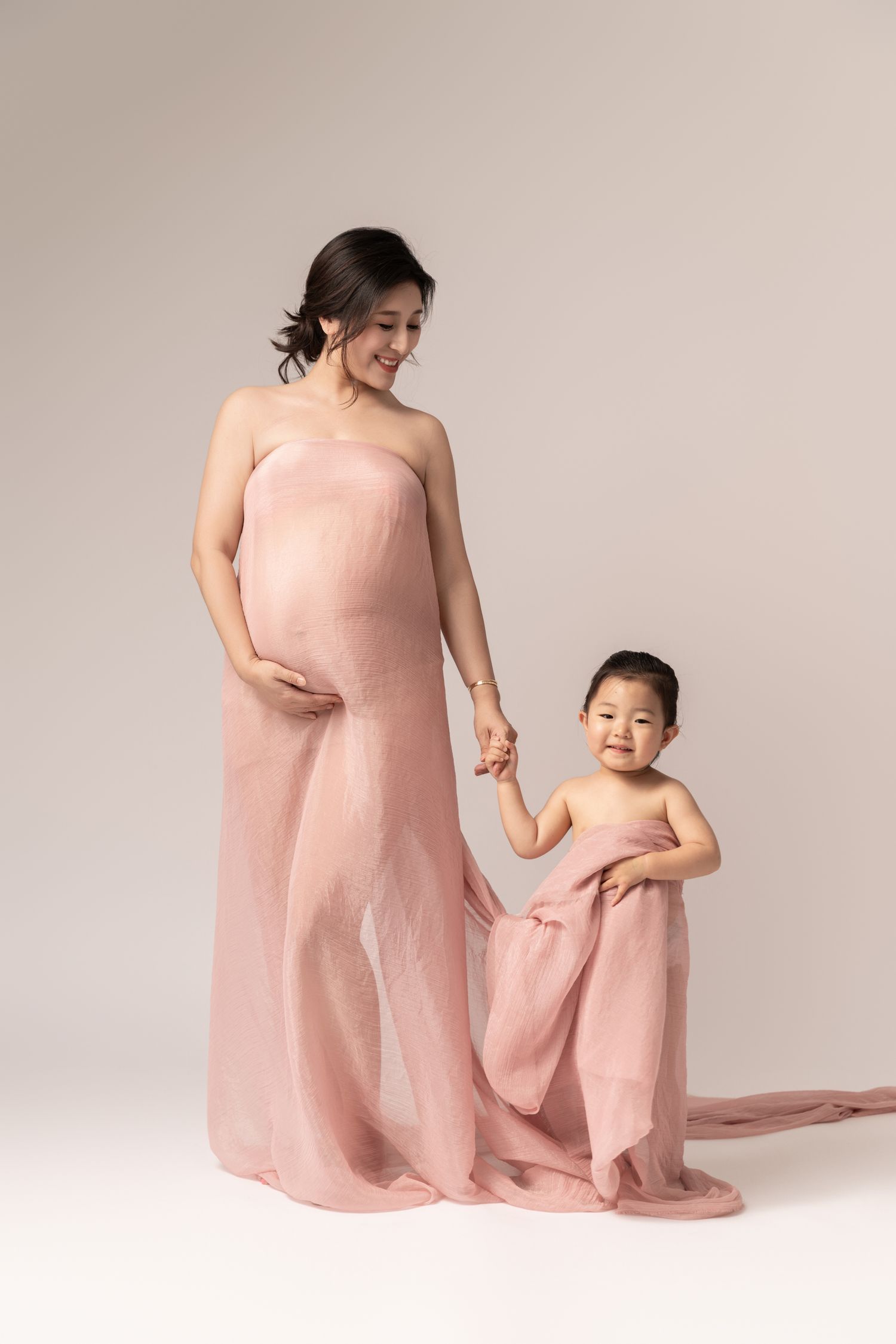 Two figures in soft pink dresses share tender moments in minimalist studio setting.