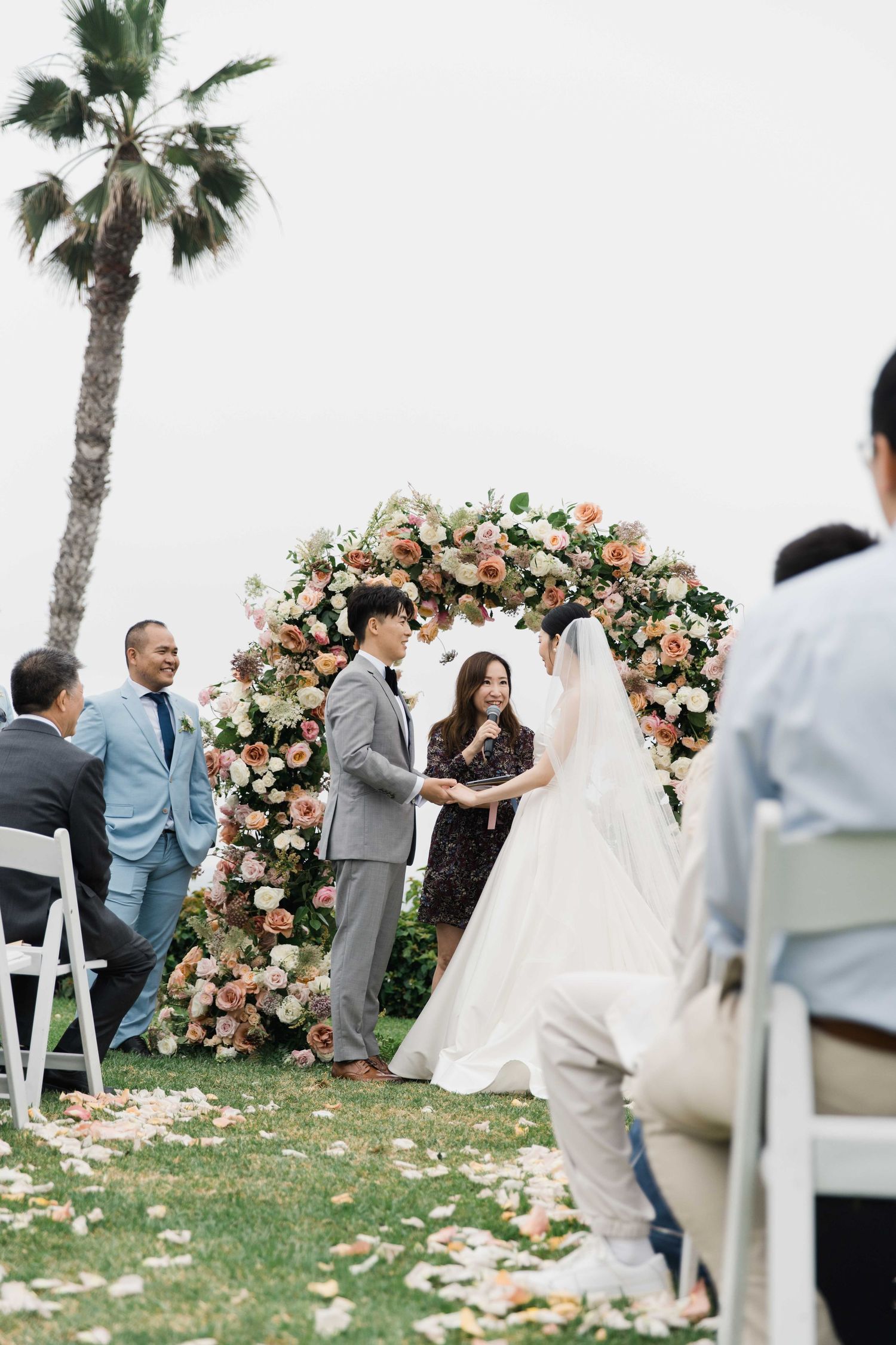 Wedding ceremony under a lush floral arch with pink and peach blooms next to a palm tree.