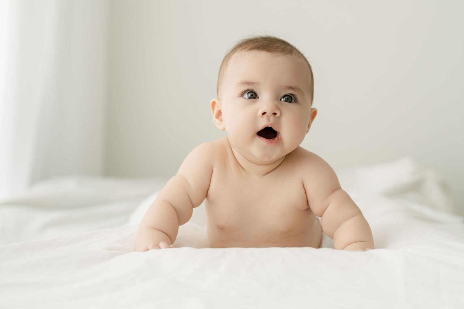 Baby with bright eyes lying on white bedding in natural light.