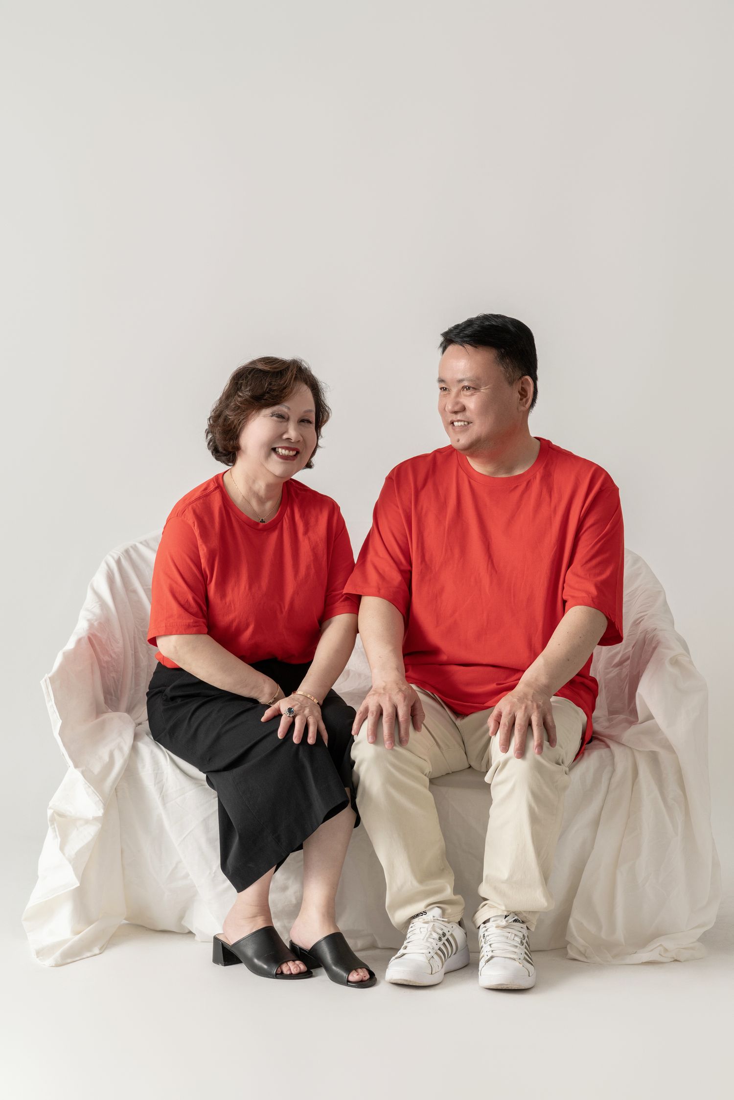 Couple in red and casual wear sits together on white studio furniture for portraits.