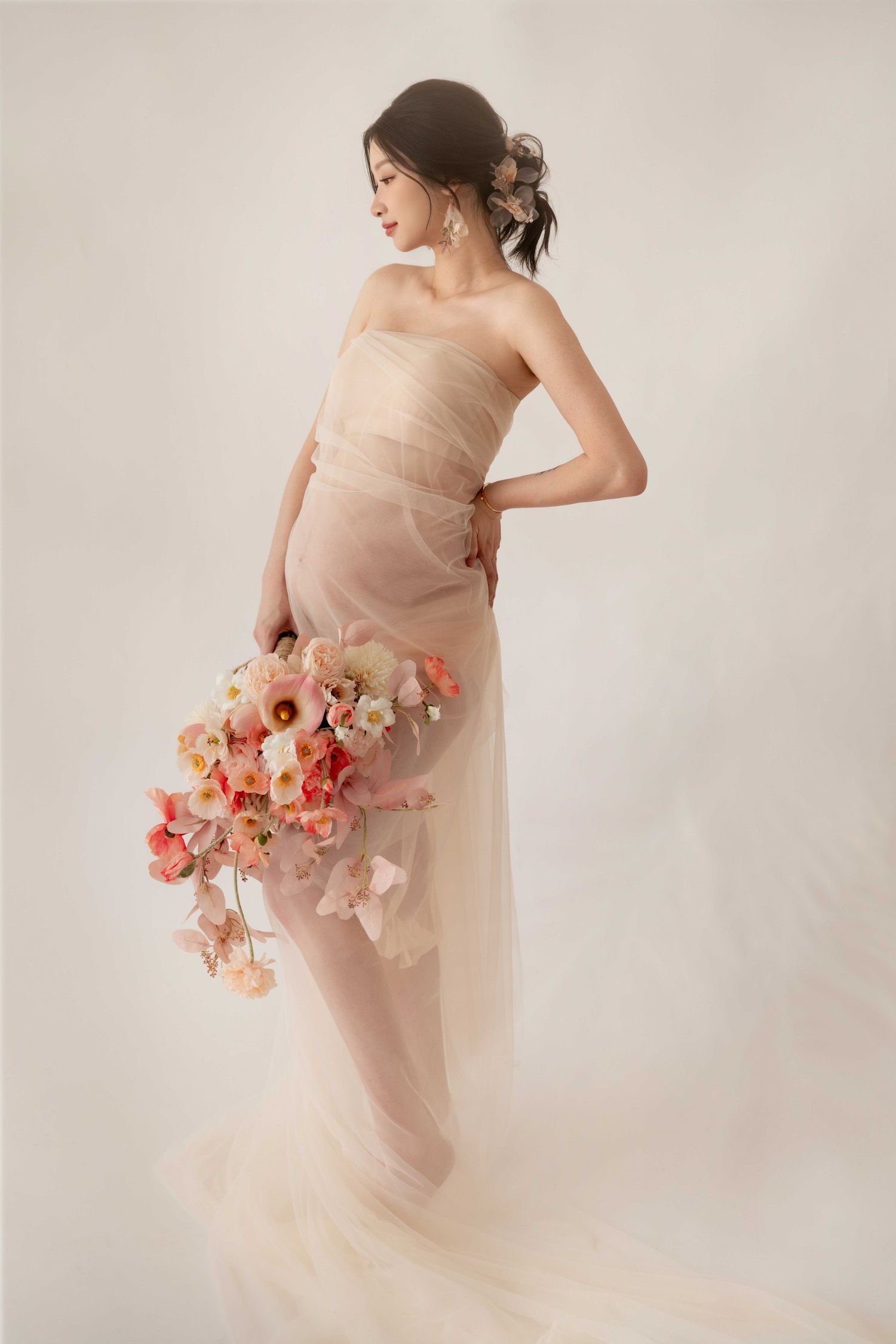 Elegant maternity portrait with coral flowers and sheer blush dress against neutral backdrop.
