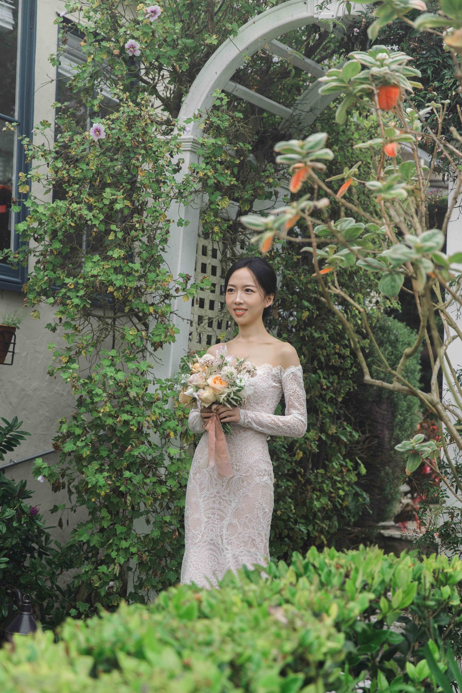 A bride holding a bouquet poses in a garden setting with blooming flowers and greenery.