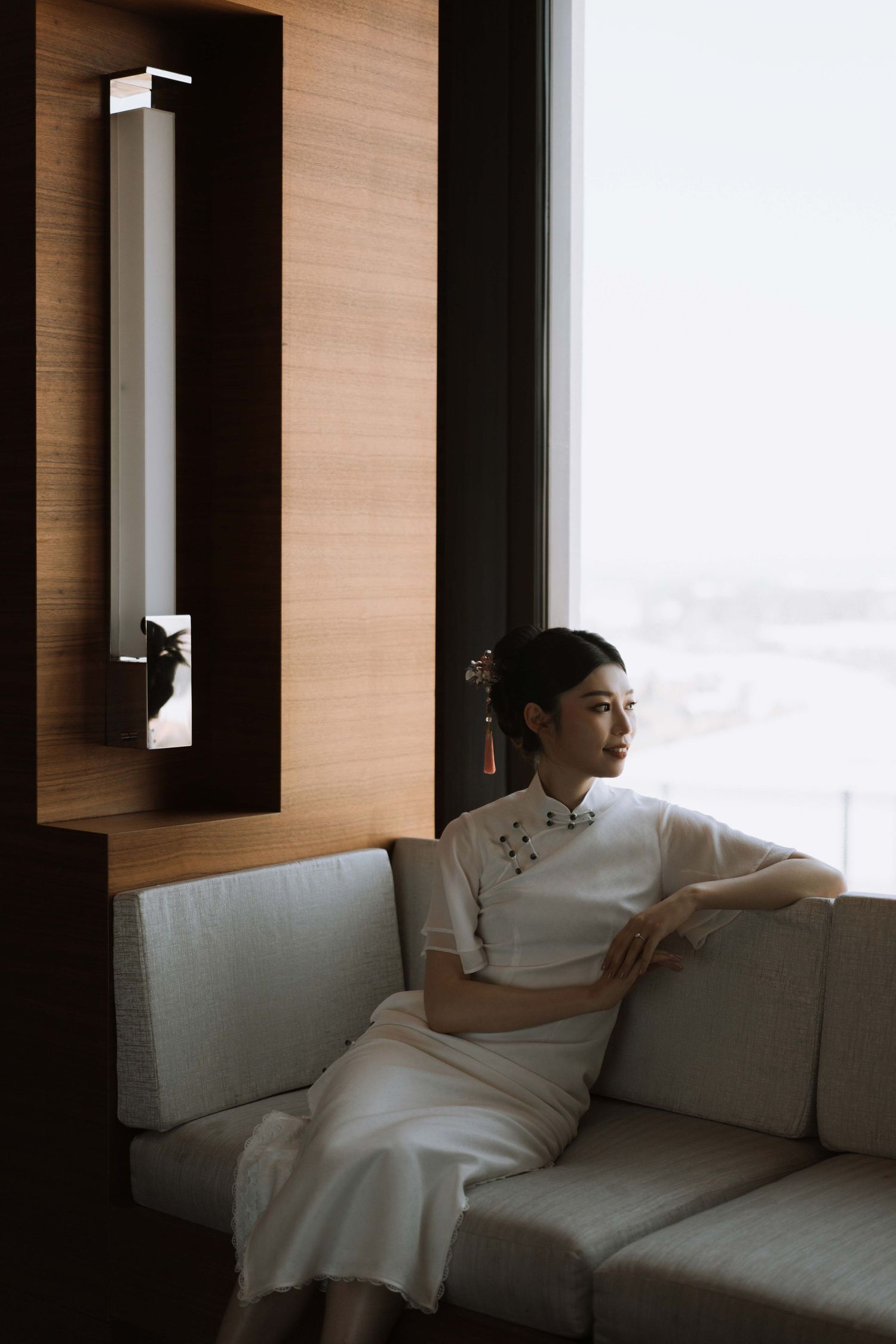 Artistic wedding photography of someone in white dress seated on modern white couch near window.