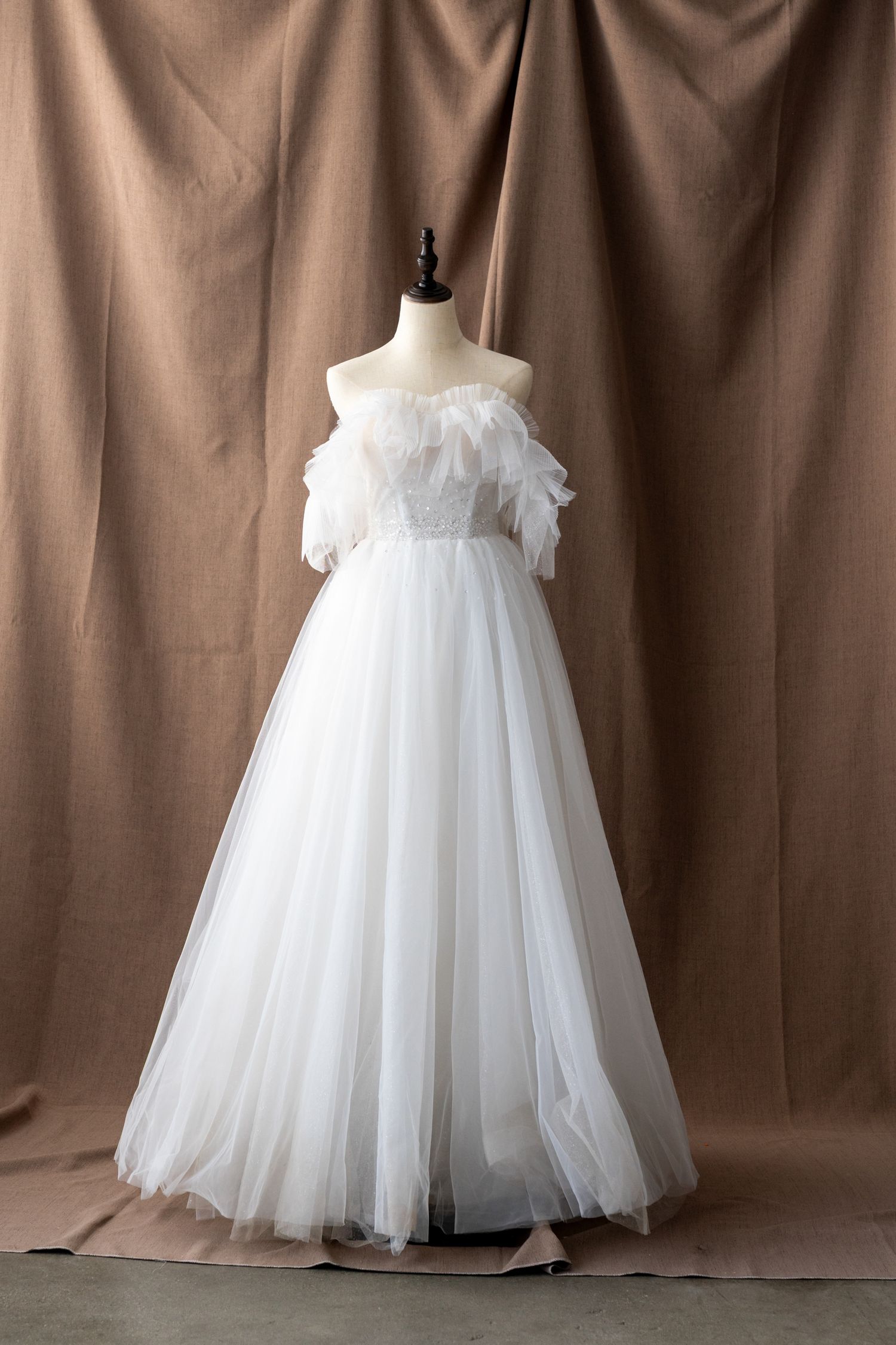 Elegant off-shoulder white tulle wedding gown with feathered bodice displayed on mannequin against brown backdrop.