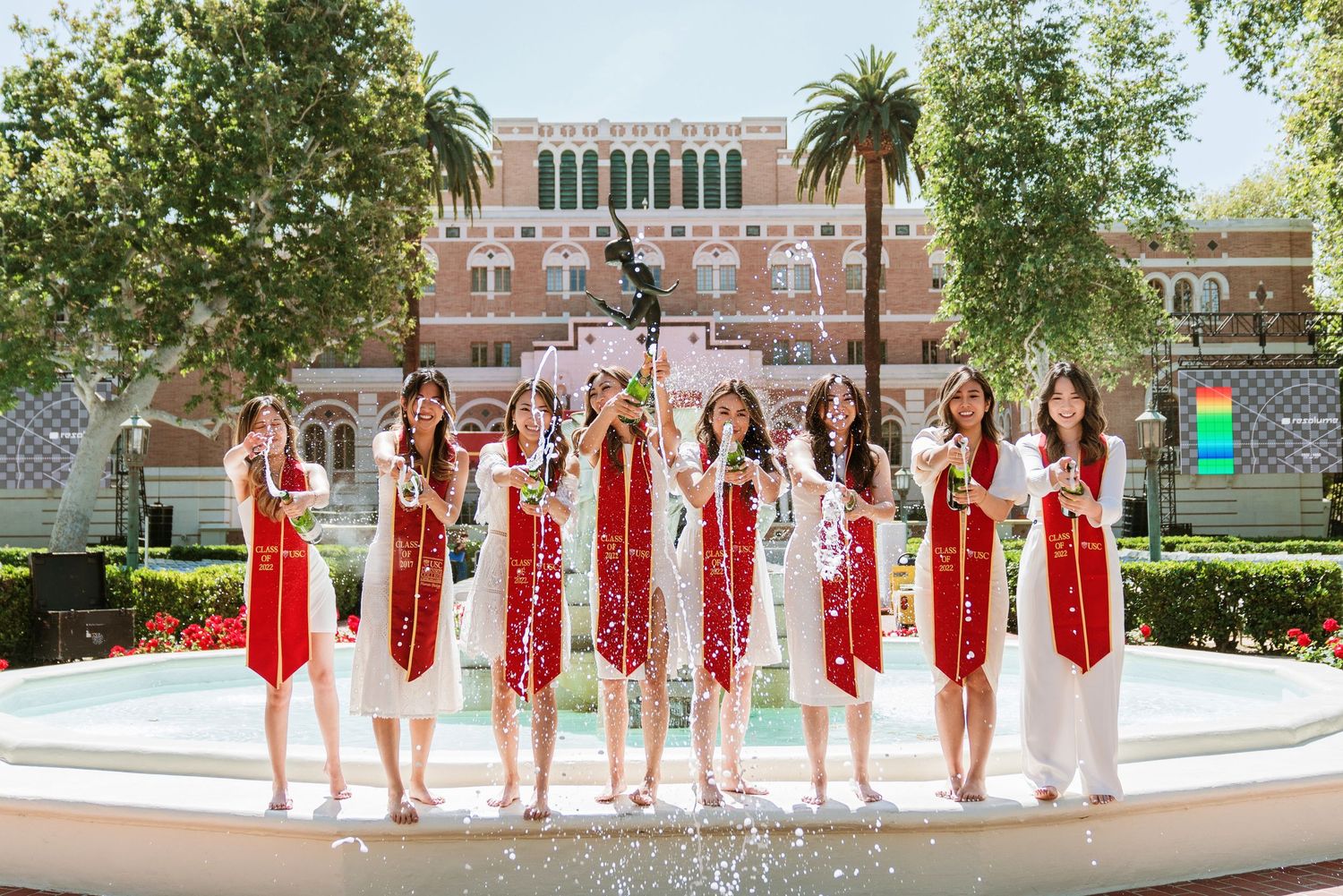 Graduates in red stoles line up for group photo on university campus with palm trees.