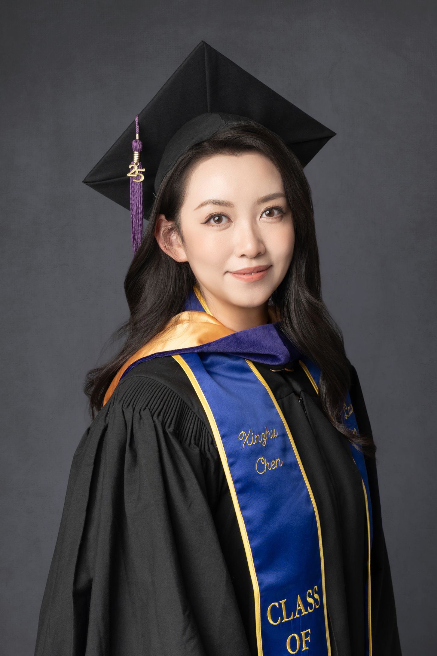 Graduate in black academic regalia with blue and gold graduation stole against dark backdrop.