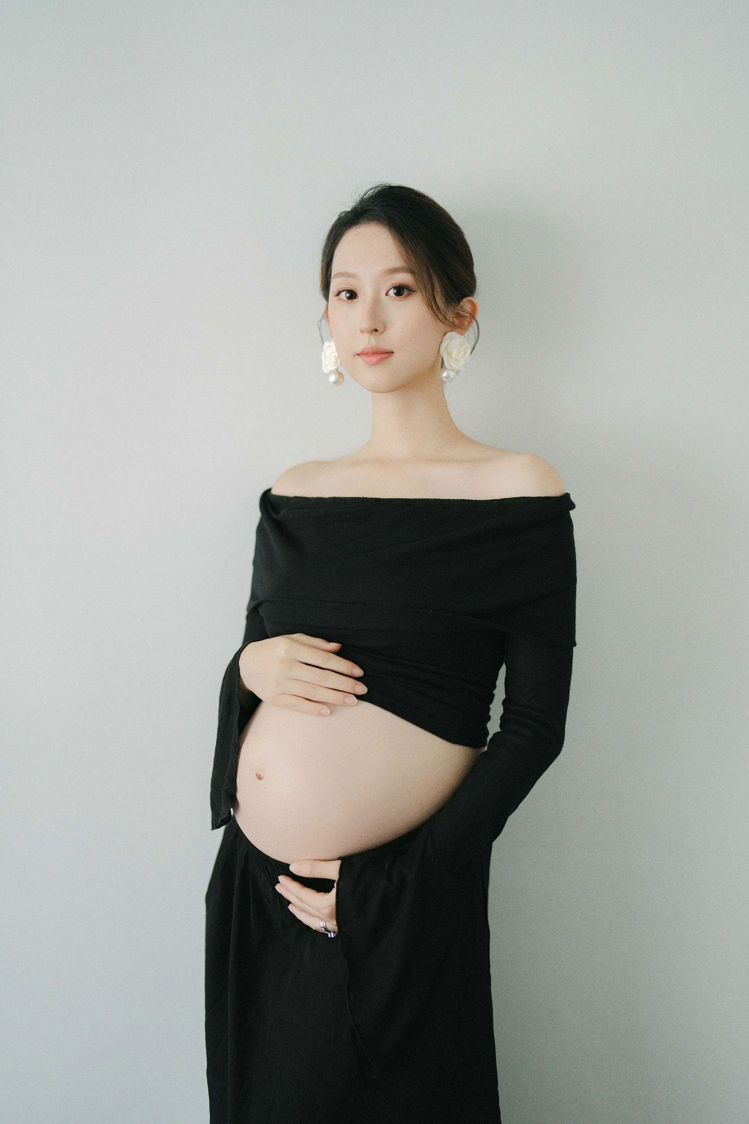 Elegant maternity photo of woman in black off-shoulder dress against white background.