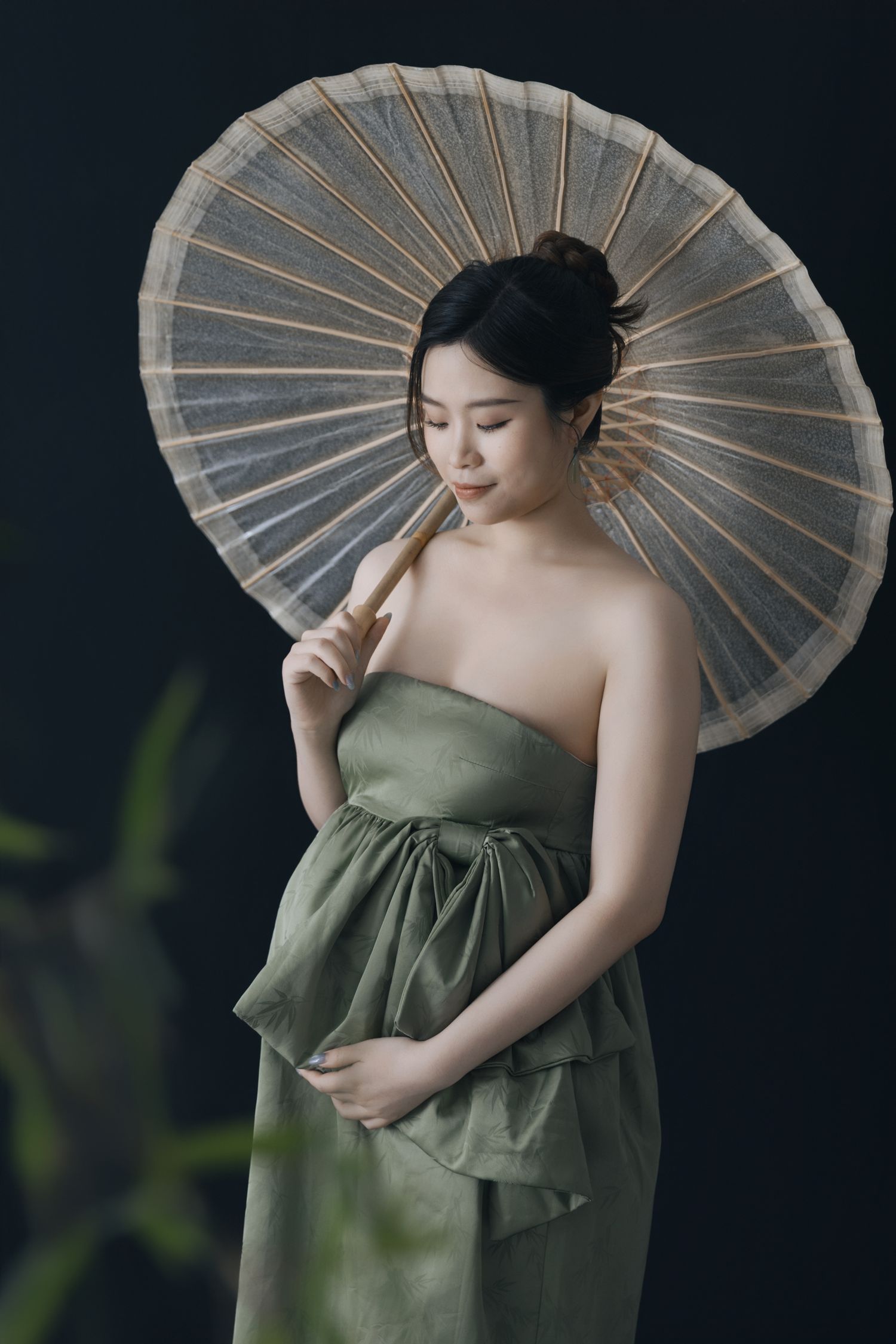 Artistic maternity photoshoot featuring a sage green dress and paper parasol against dark background.