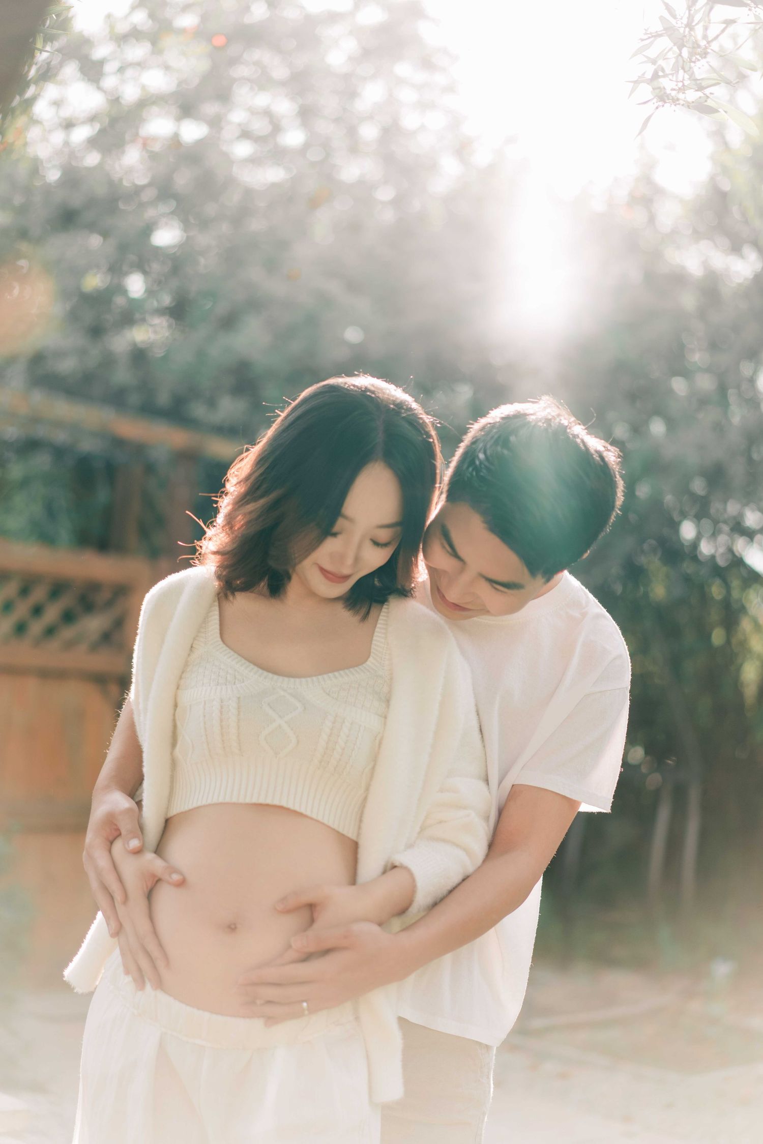 Sun-drenched maternity photos of couple in white outfits embracing outdoors.