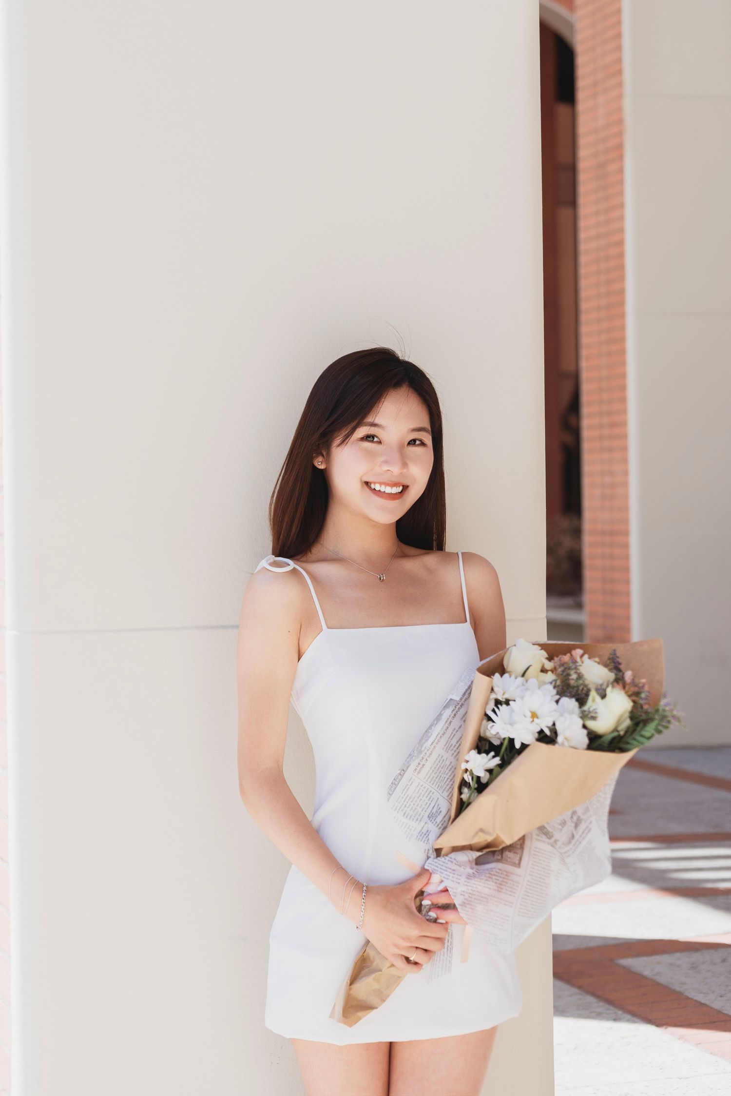 Graduate in white dress holding flowers poses in columned hallway.