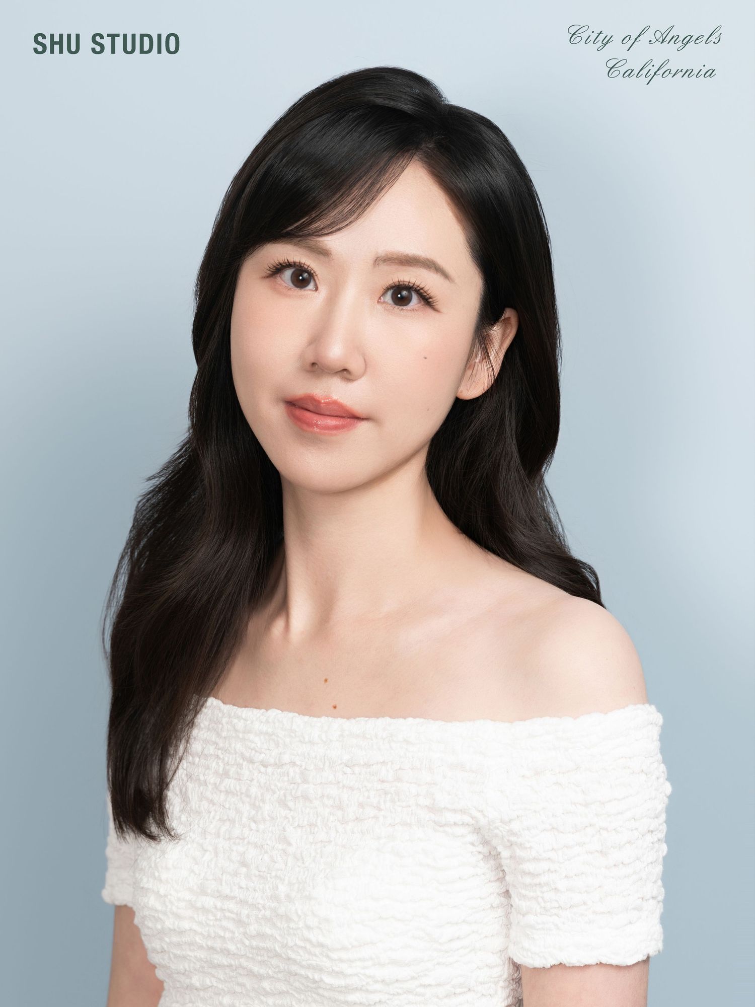 Studio portrait in white off-shoulder textured top against light blue background.