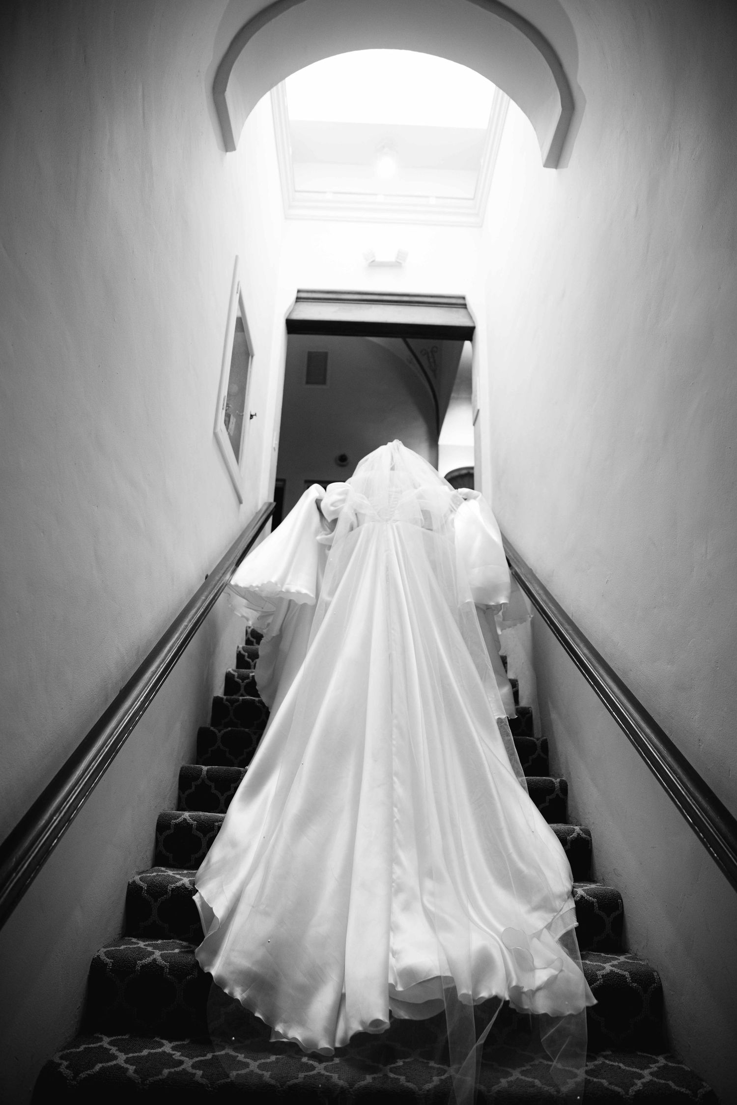 A dramatic black and white photo of a wedding dress descending a narrow staircase.
