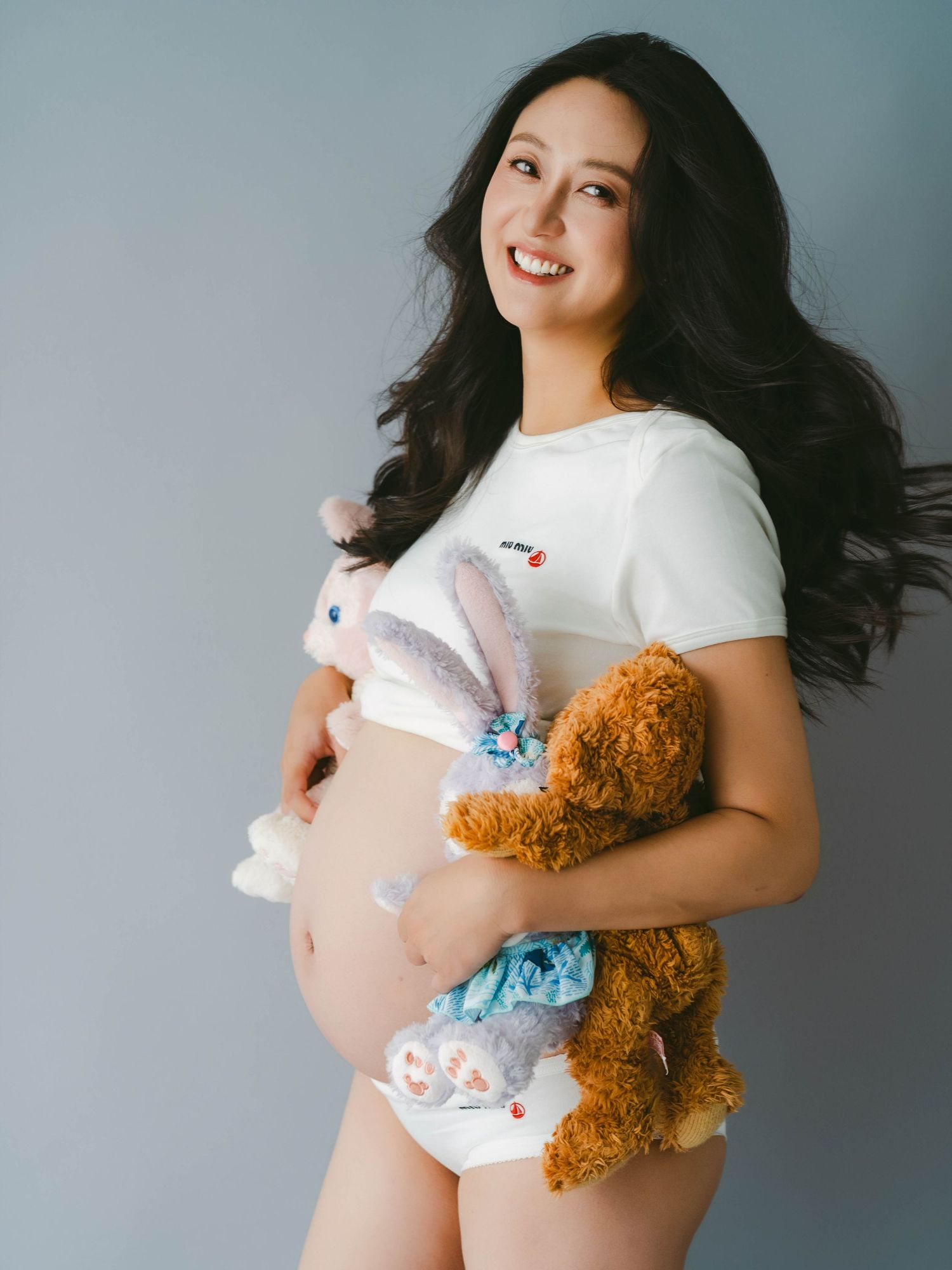 Casual maternity photo with teddy bear and white shirt against gray backdrop.