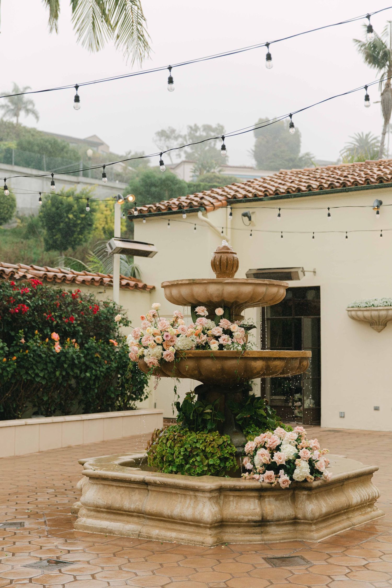 Stone fountain decorated with peach and white flowers on terracotta patio with string lights overhead.