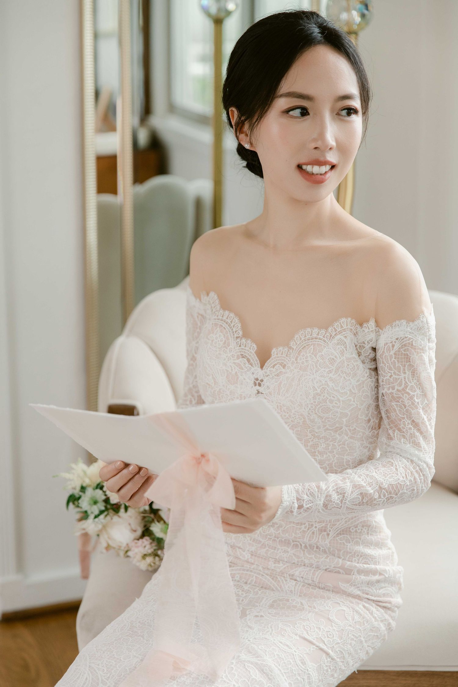 Bride in white lace dress sits elegantly while reading in a sunlit room with classic decor.