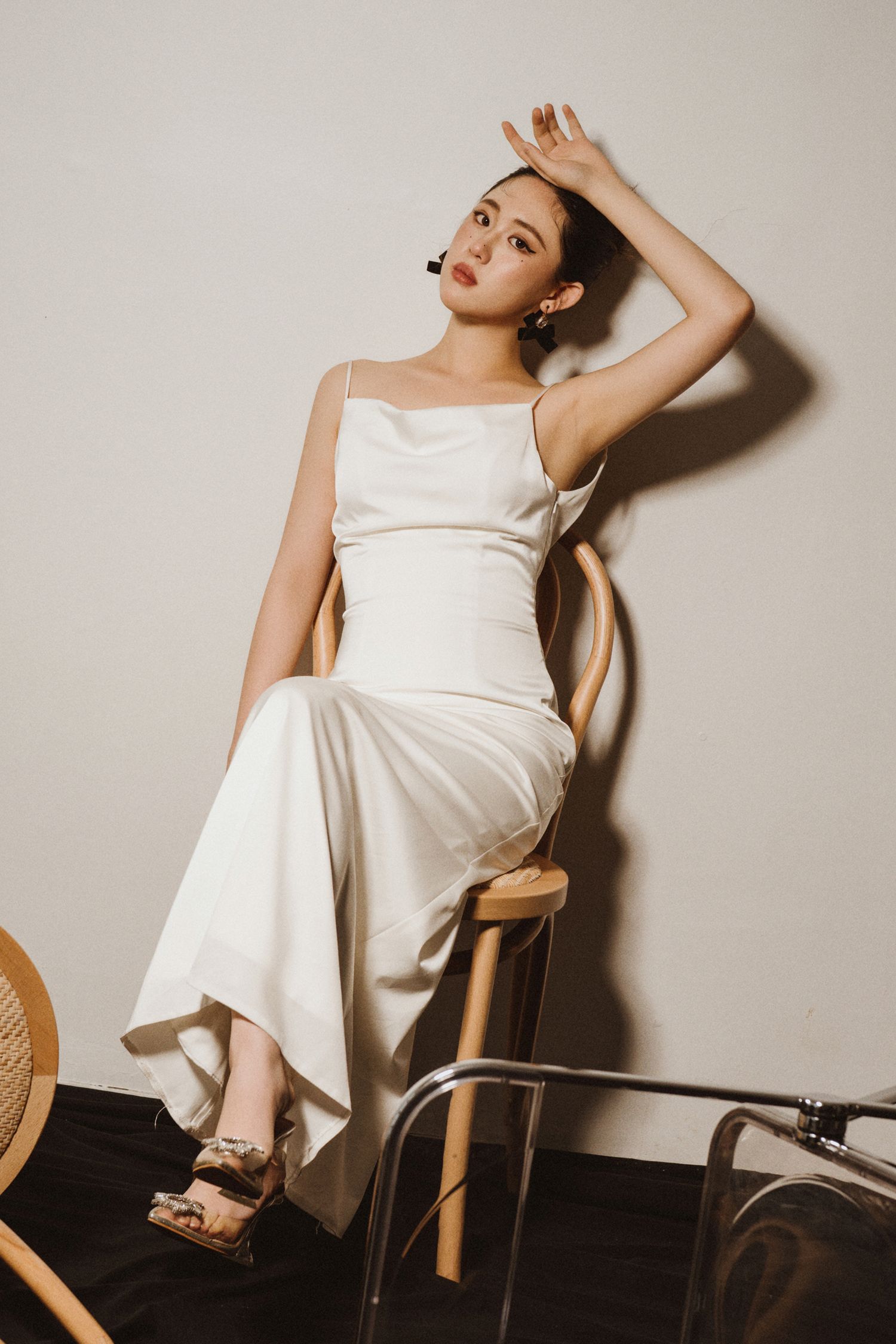 Fashion model in white dress poses artistically on wooden chair against neutral backdrop.