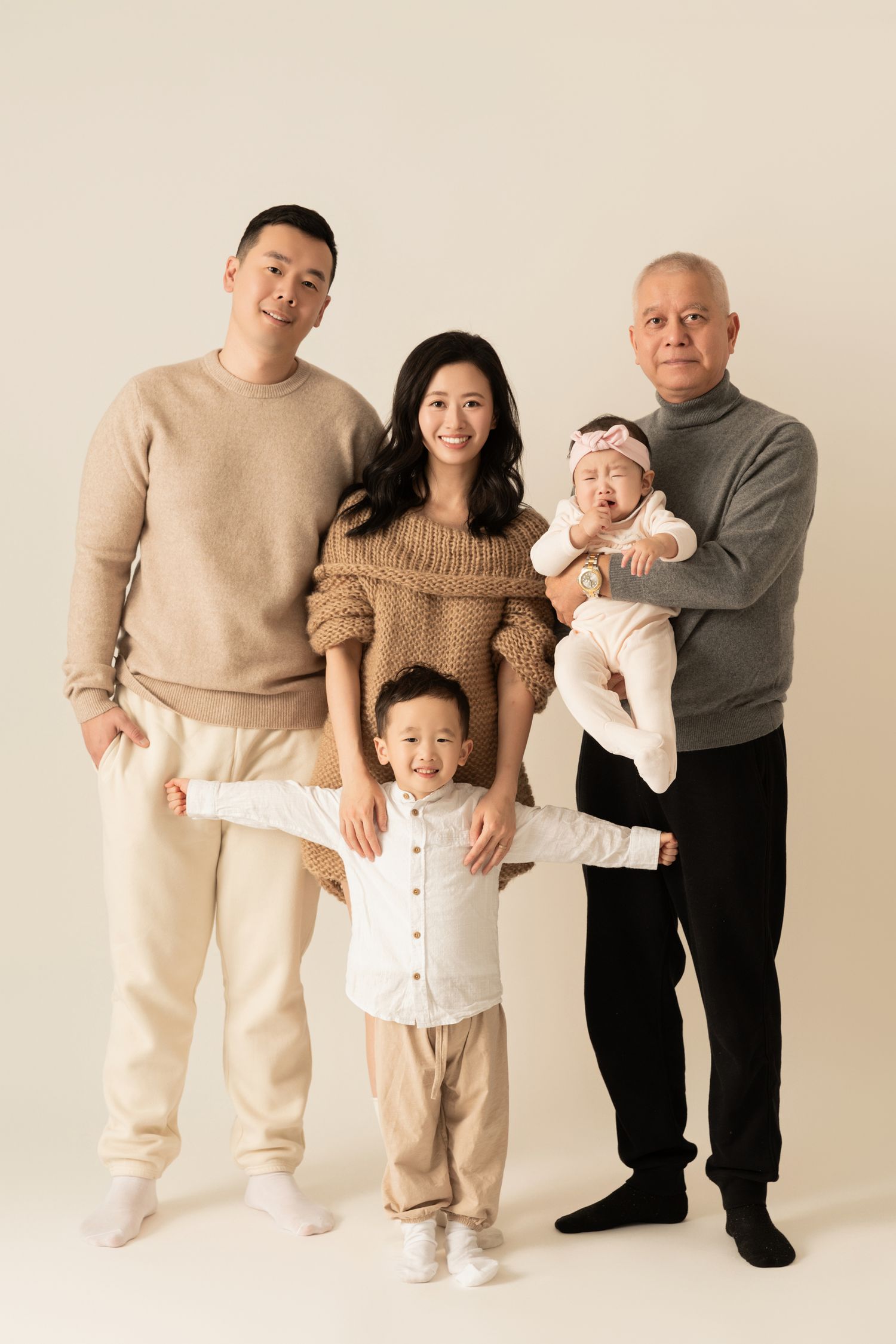 Three generations pose together in coordinated neutral outfits against beige backdrop.