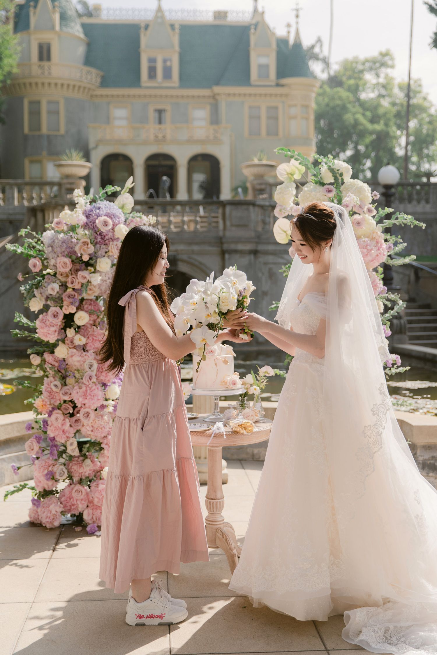 Bridesmaids in pink dresses gather around a beautiful white wedding gown in an elegant outdoor setting.