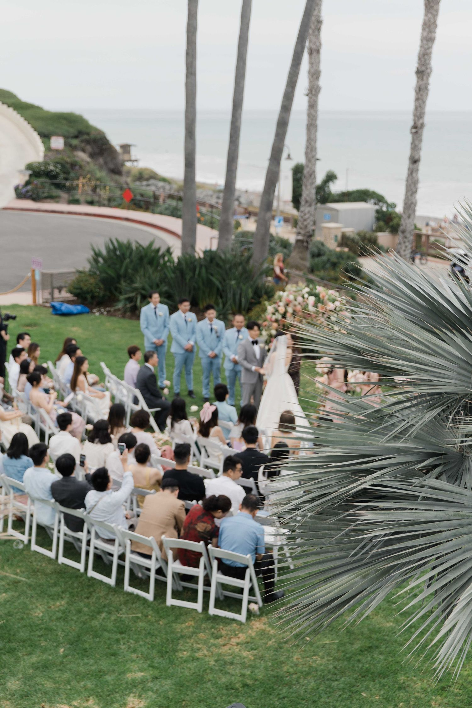 Aerial view of a beachside wedding ceremony with palm trees and white chairs on a grassy lawn.