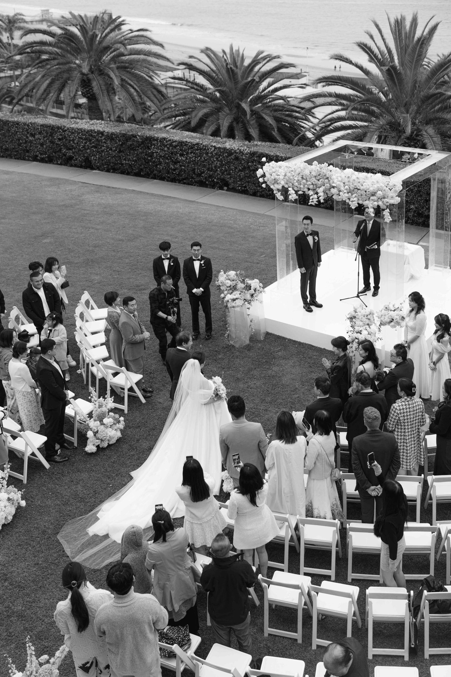 Outdoor wedding ceremony setup with white chairs and floral arrangements under palm trees.