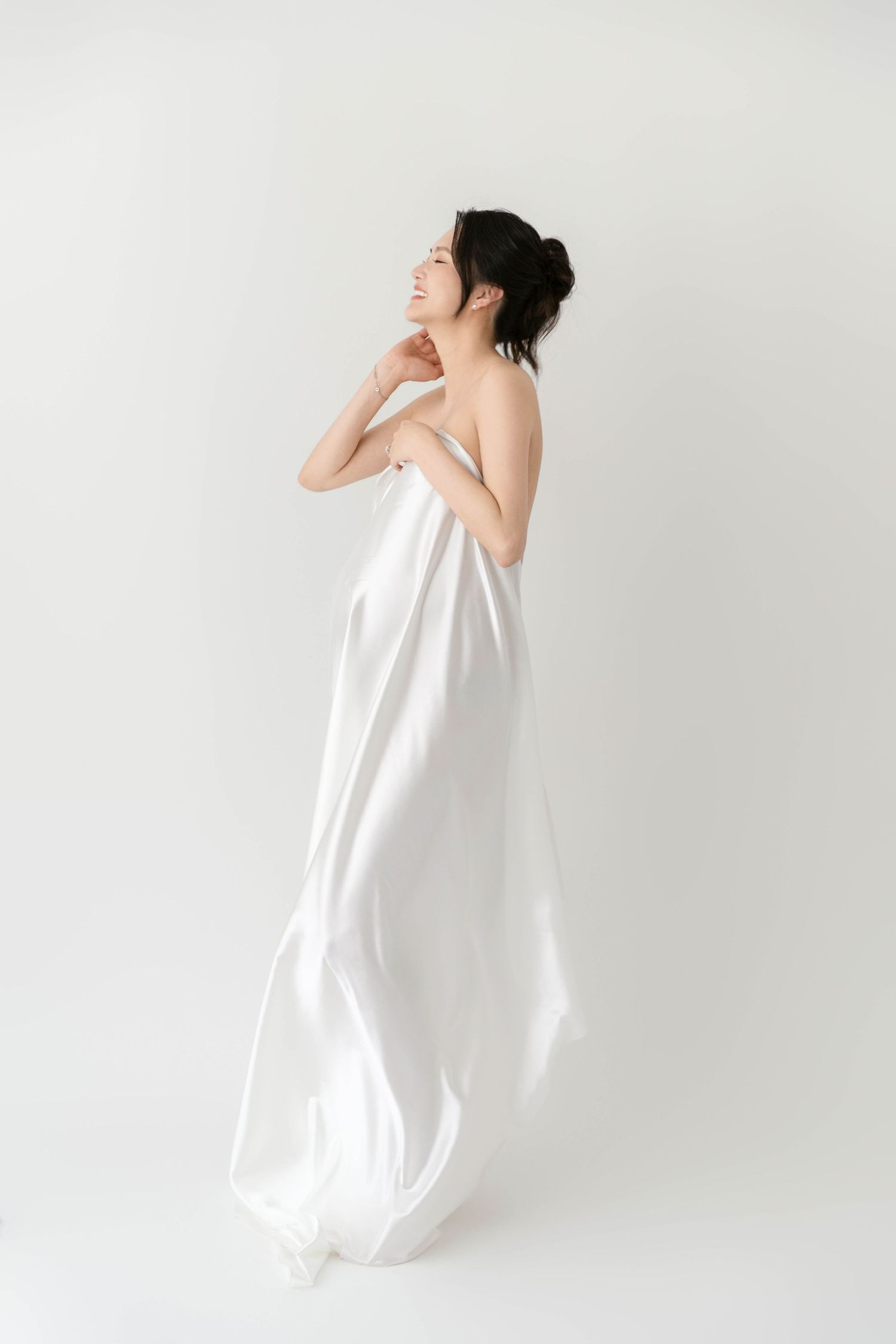 Elegant maternity pose with flowing white fabric in studio setting.
