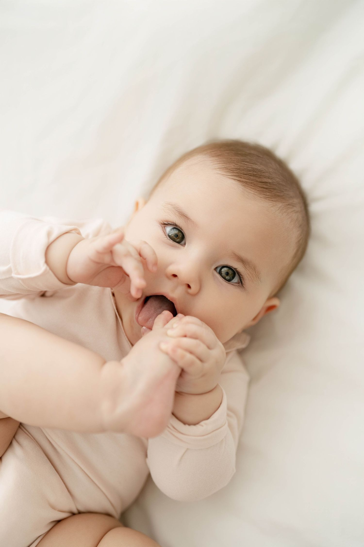Baby in light colored onesie playing with hands on white bedding.