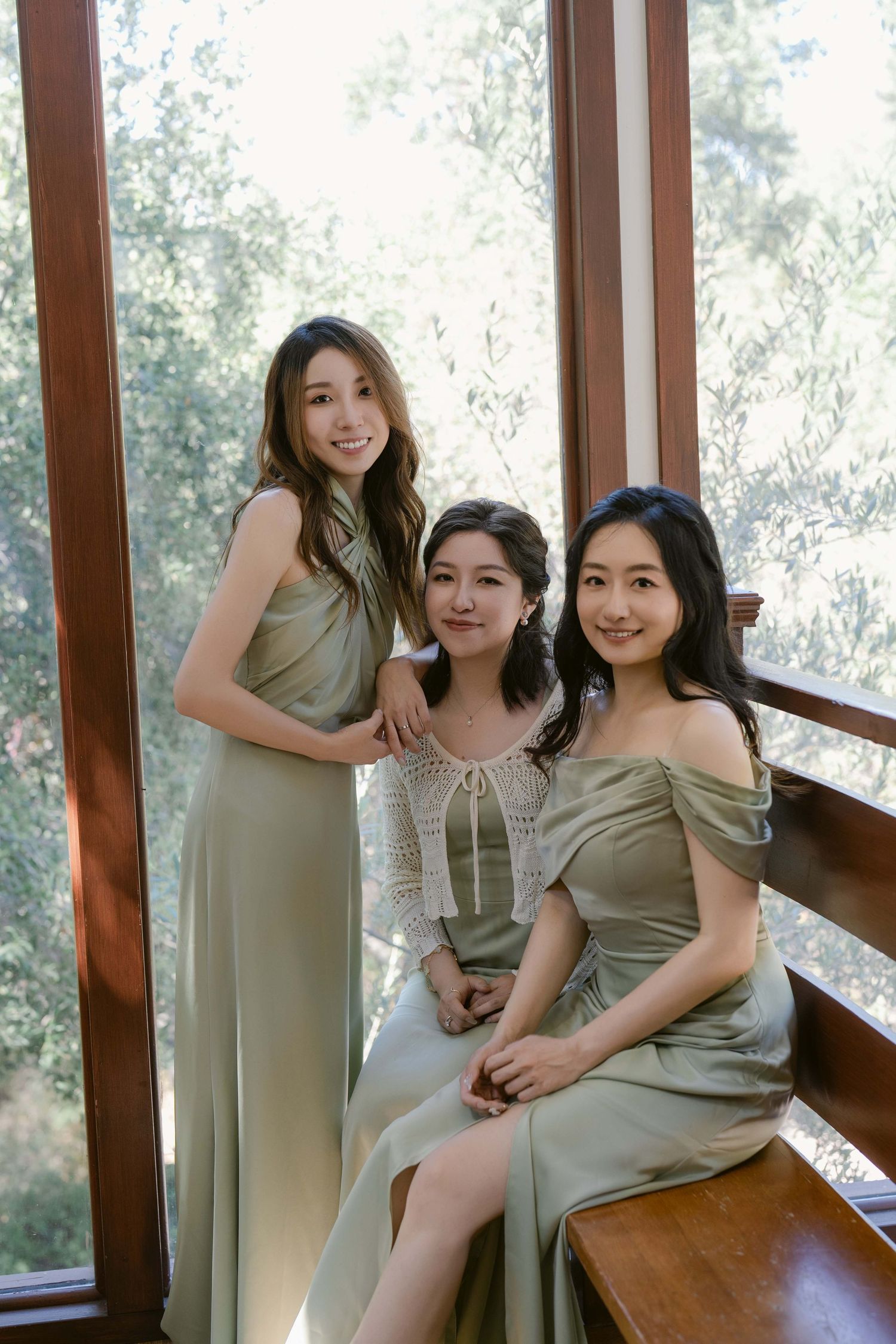 Bridesmaids in sage green dresses pose together on a wooden bench near large windows.