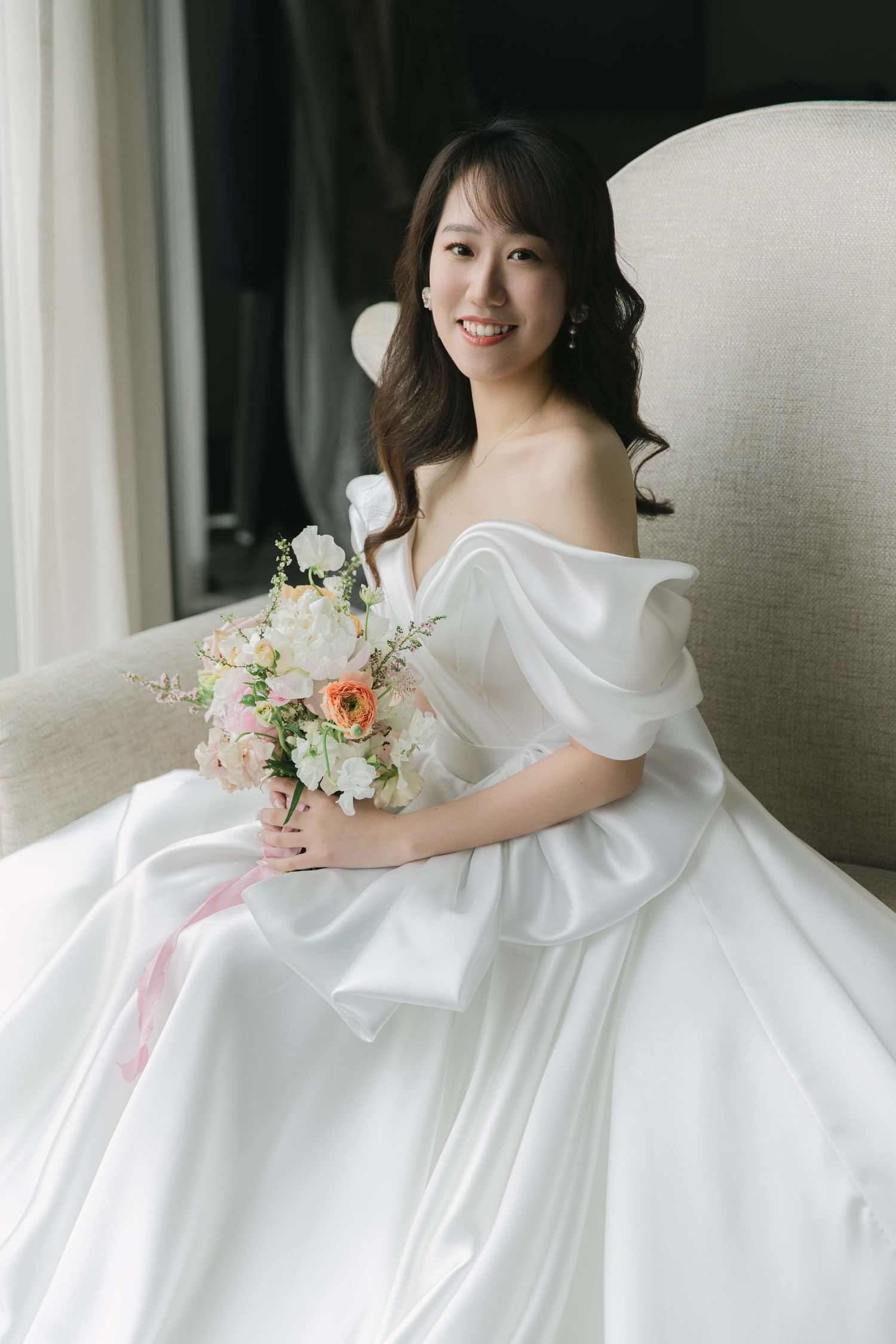 Elegant bride in white satin gown holds delicate peach and white floral bouquet against light background.