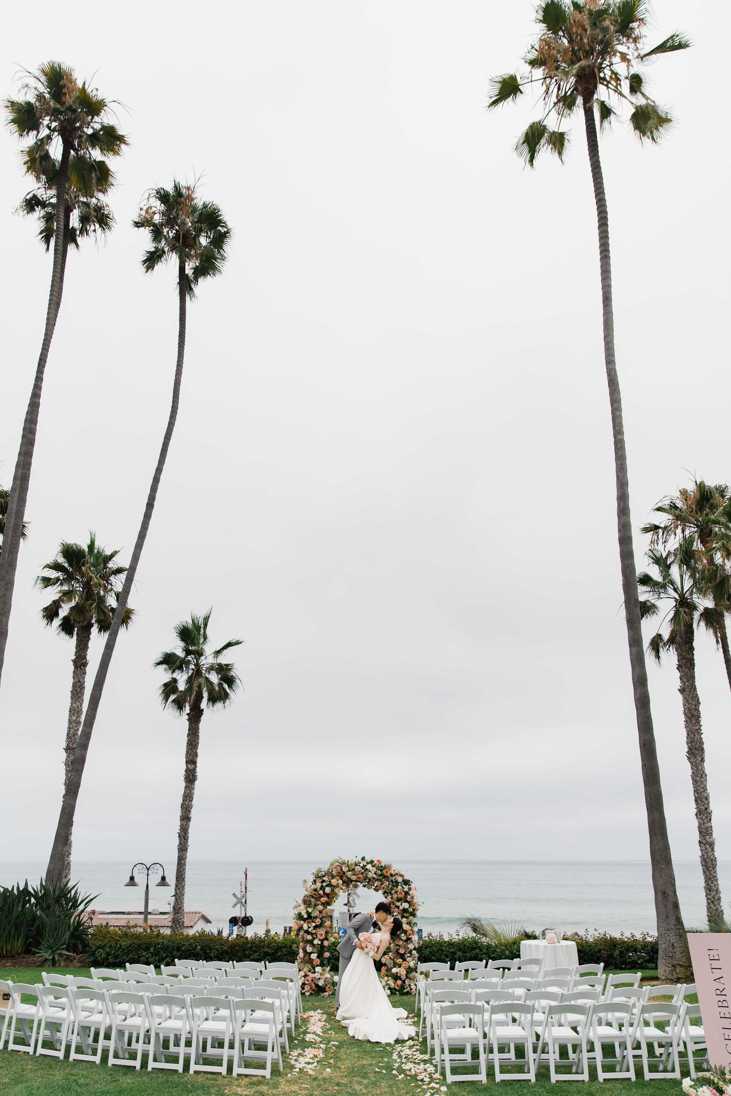 Palm tree-lined wedding venue with white chairs arranged in rows facing a floral ceremony arch.