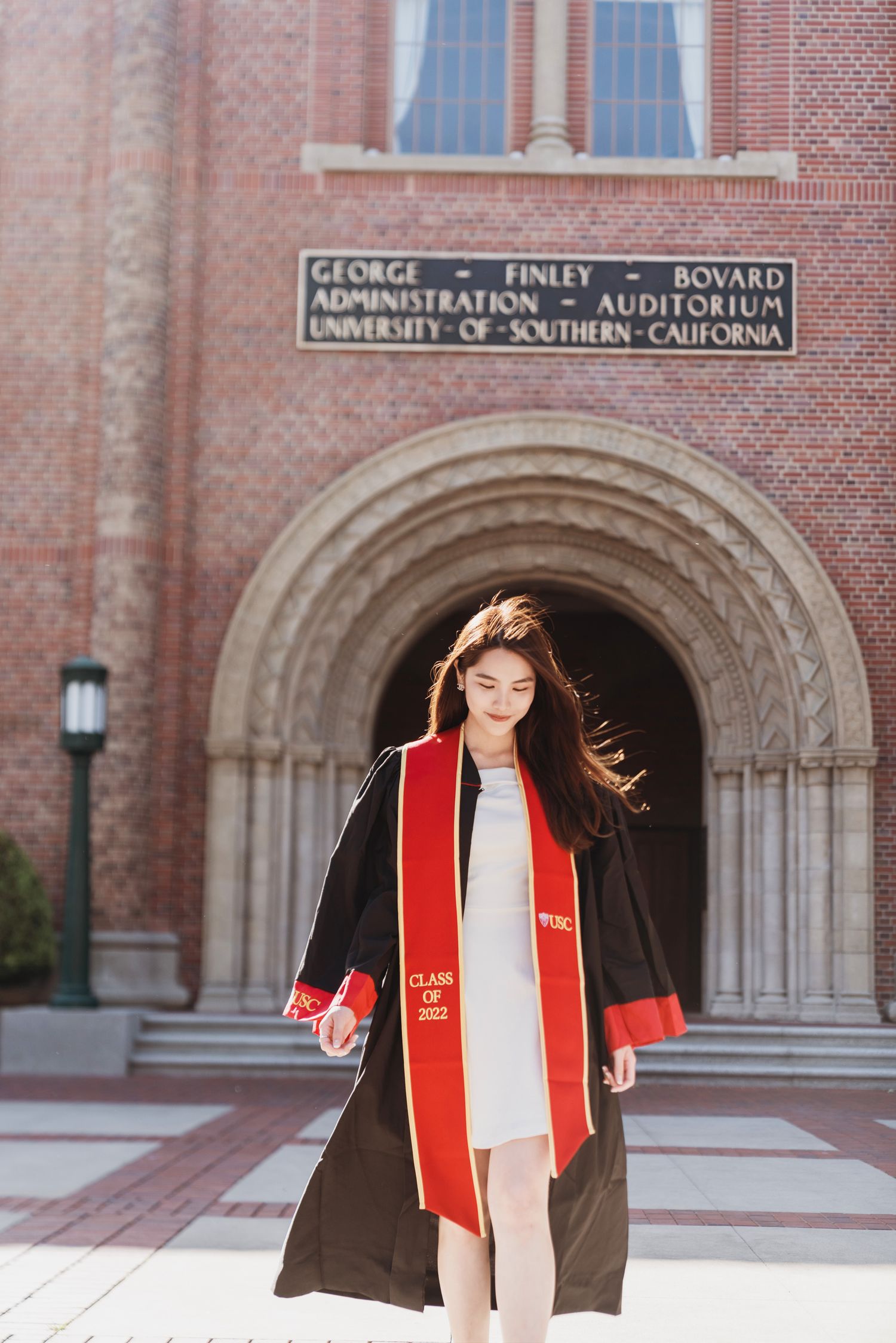 Graduate in red and black regalia poses in front of Bovard Auditorium's brick facade.