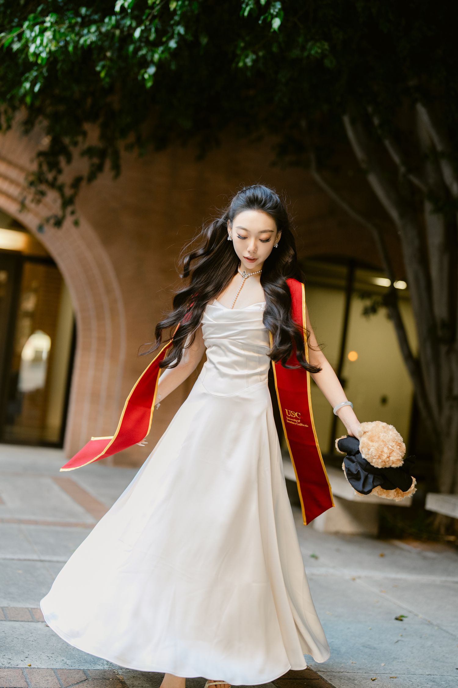 Graduate in white dress and red stole twirling on campus grounds.