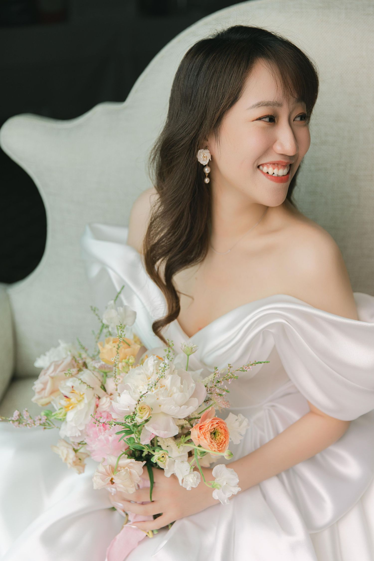 Bride in off-shoulder white dress poses with spring bouquet in natural light setting.