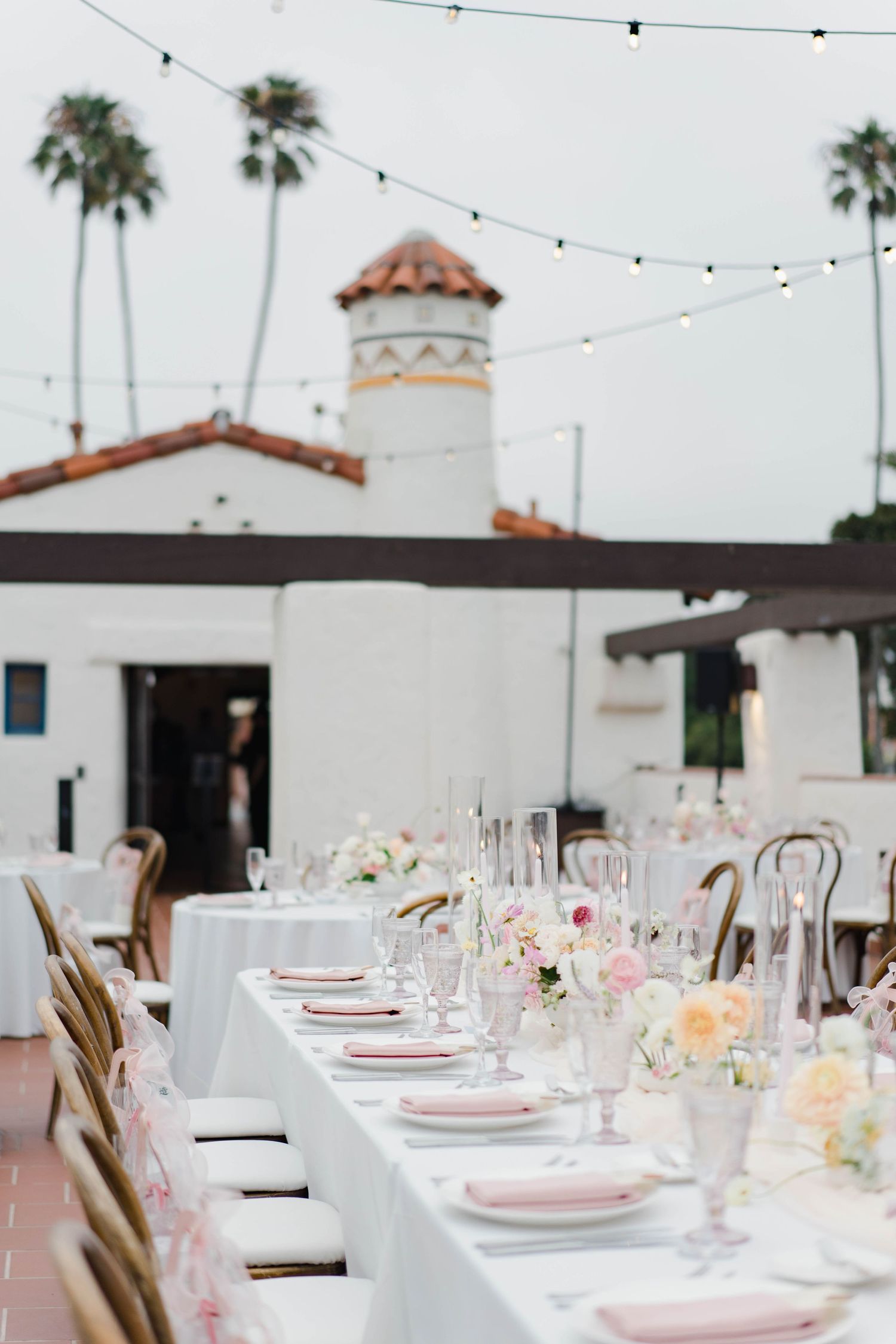 Elegant wedding reception tables set with pink accents and string lights under a Spanish-style terrace.