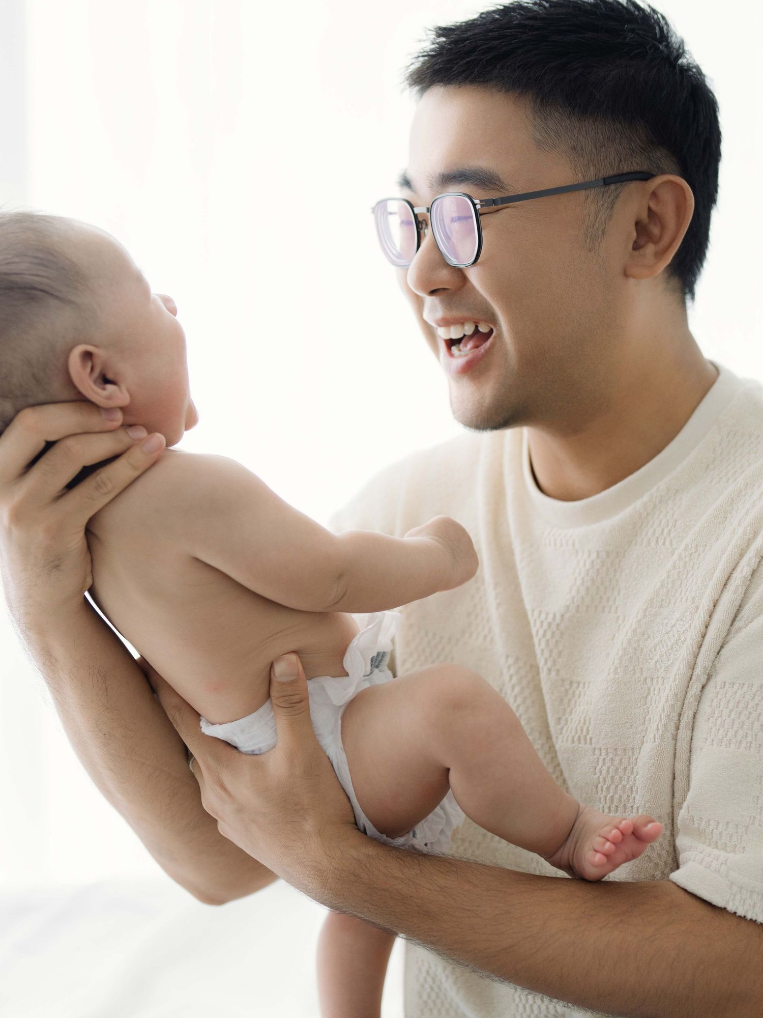 Parent playing with laughing baby in white sweater by bright window.