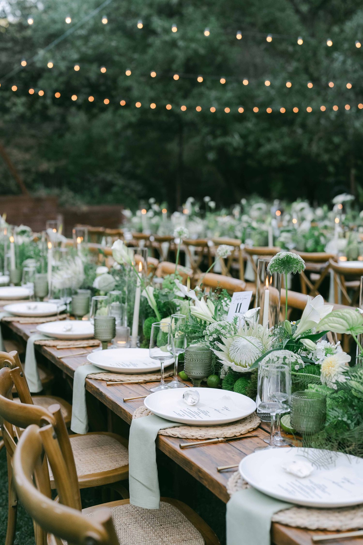 Long wooden tables set for outdoor dining with string lights and green and white decor.