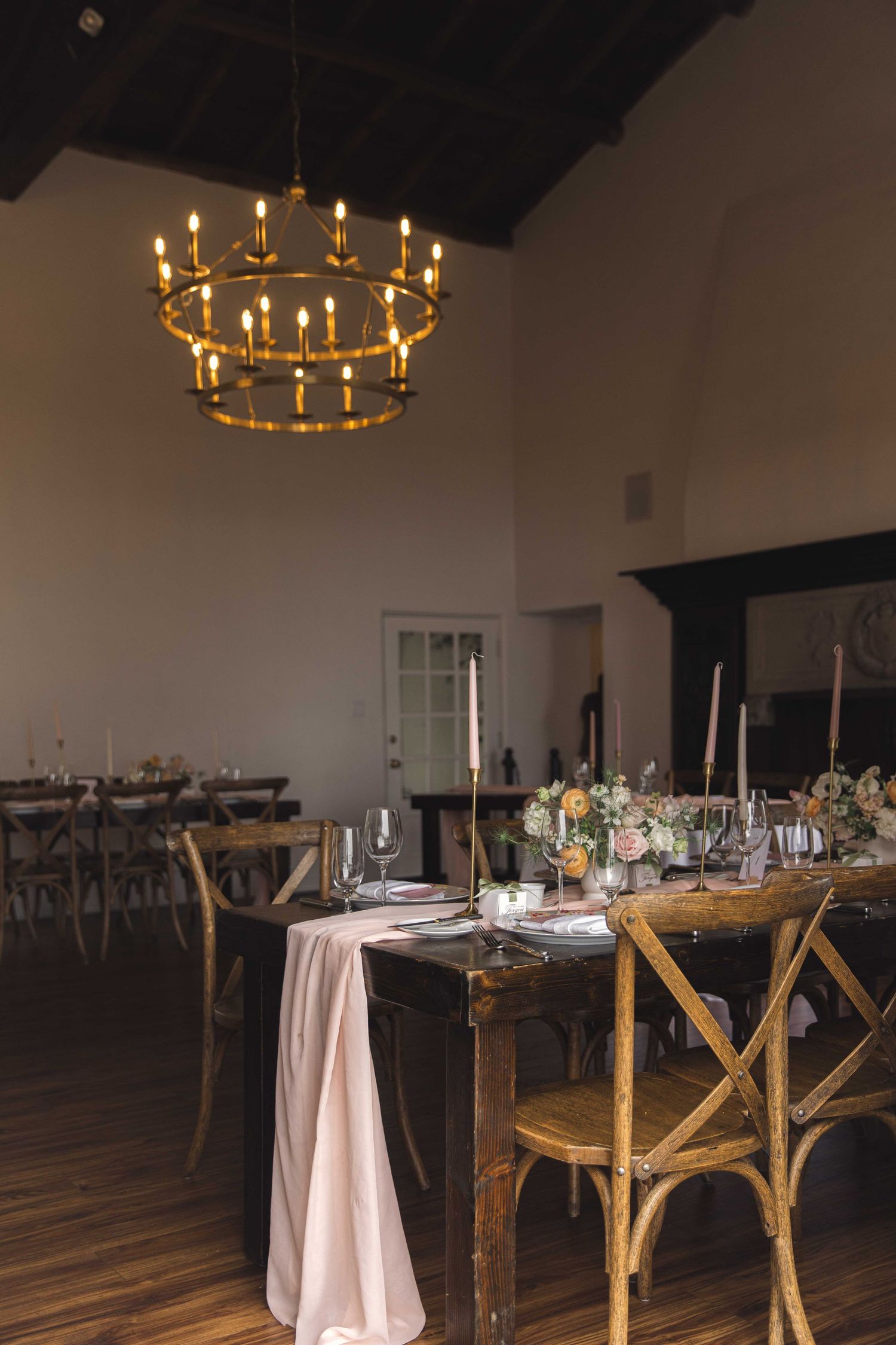 Reception venue with two-tiered brass chandelier, wooden cross-back chairs and white tablecloths.