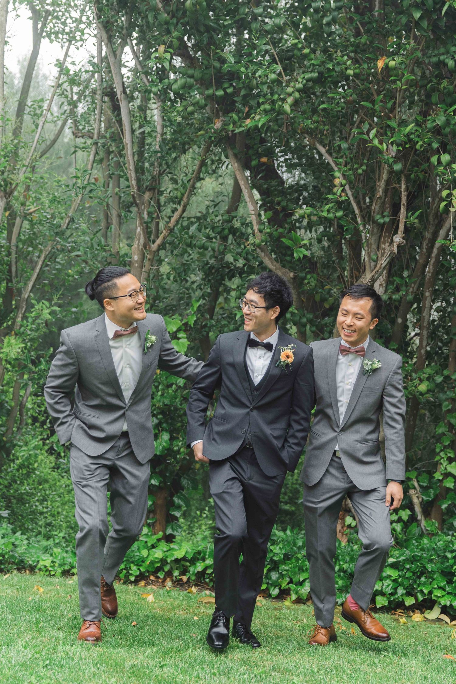 Three groomsmen in formal attire pose playfully on grass with greenery backdrop.