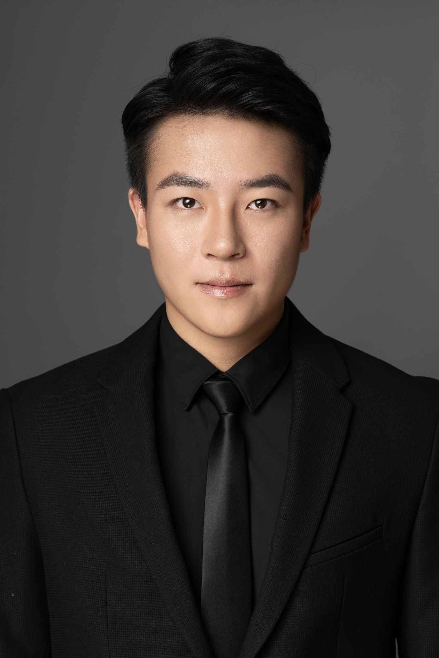 Professional portrait of person wearing black suit and tie against dark background.