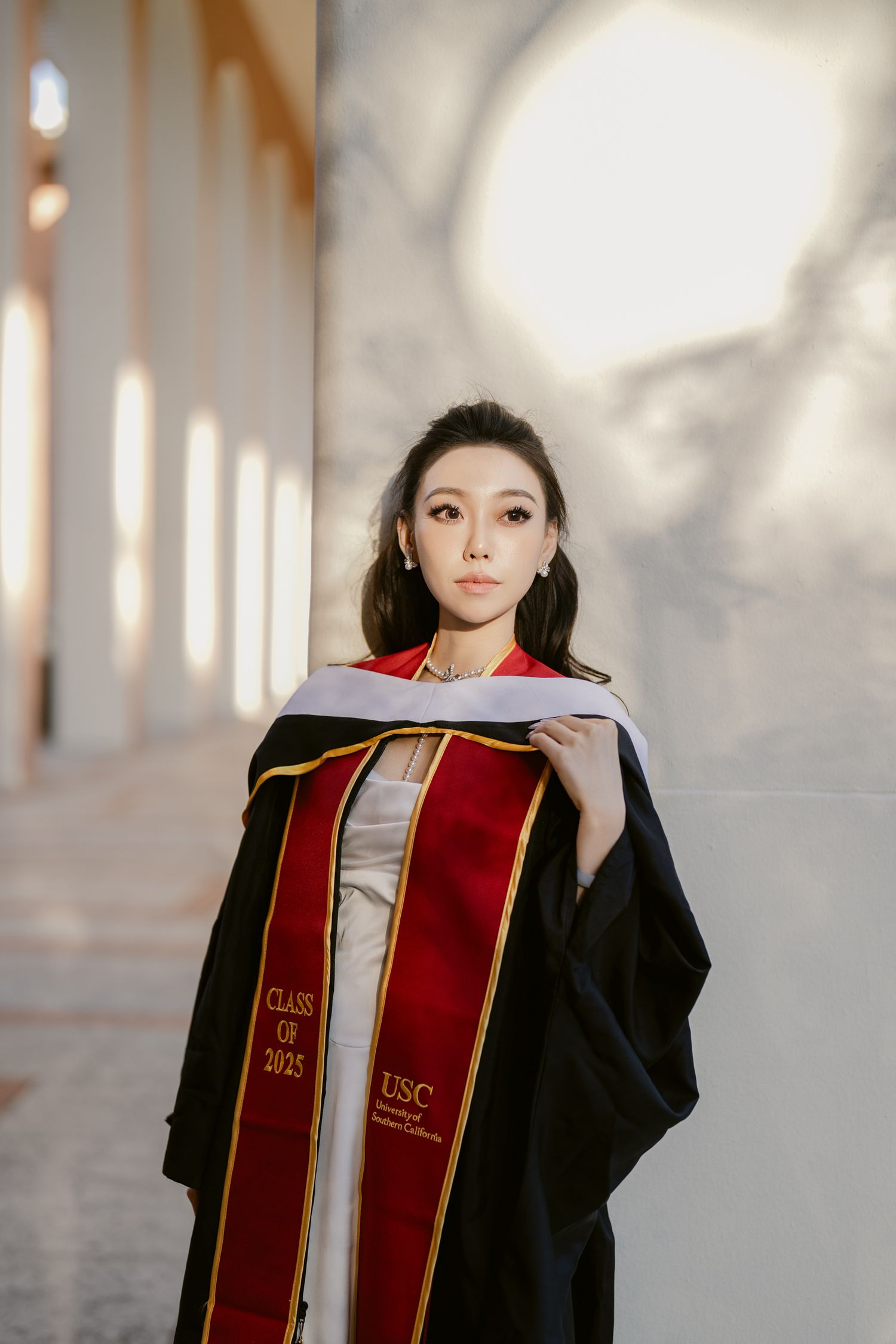 Graduate wearing black cap and gown with red stole poses in front of light-filled hallway.