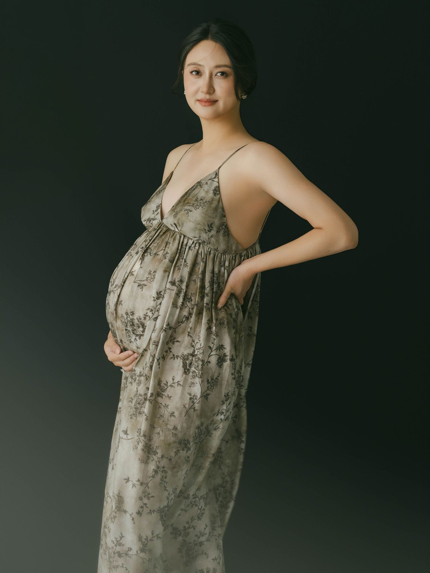 Artistic maternity portrait with woman in grey dress against dark backdrop.