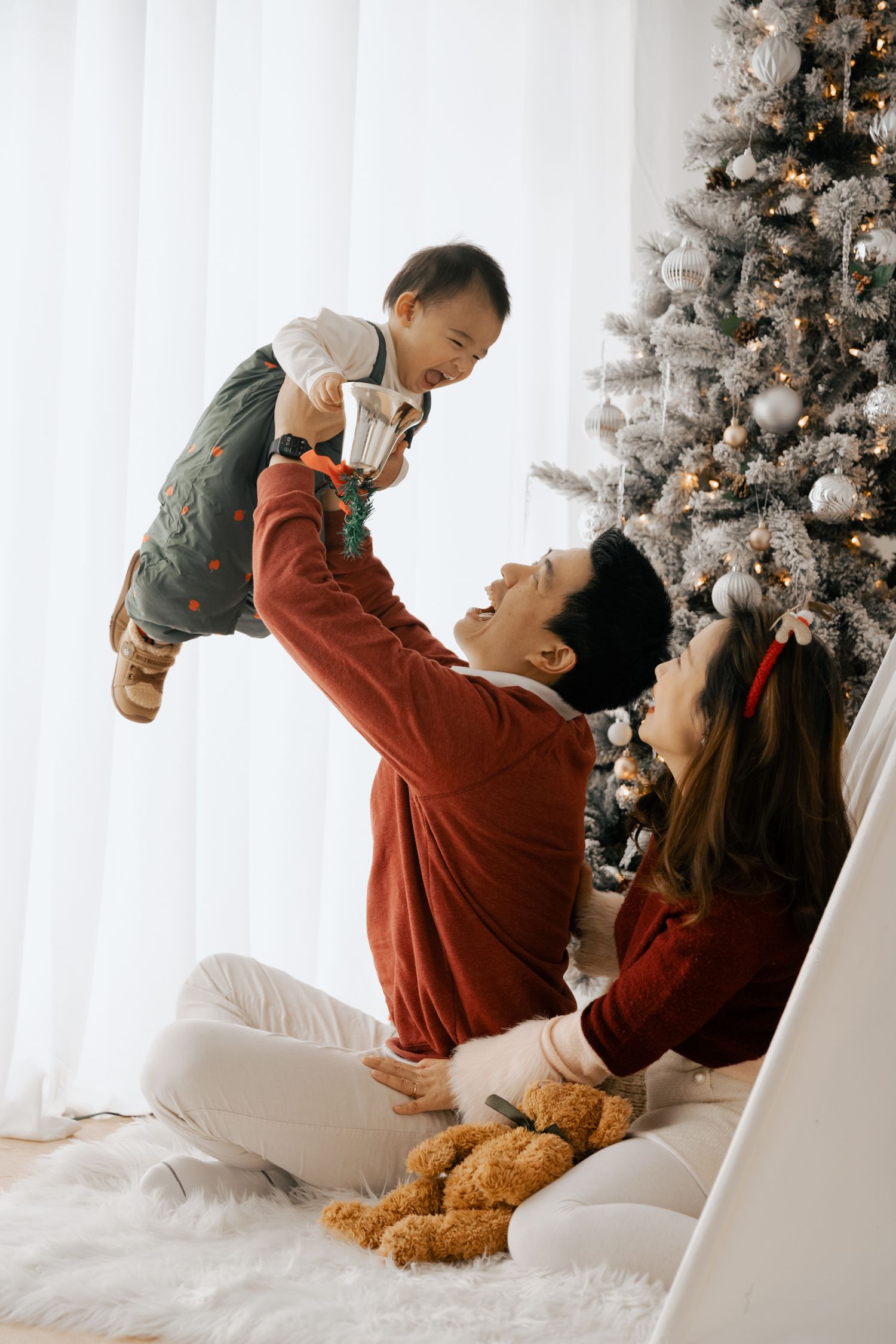 Joyful moment by Christmas tree as parents lift baby in red holiday sweaters with teddy bear nearby.
