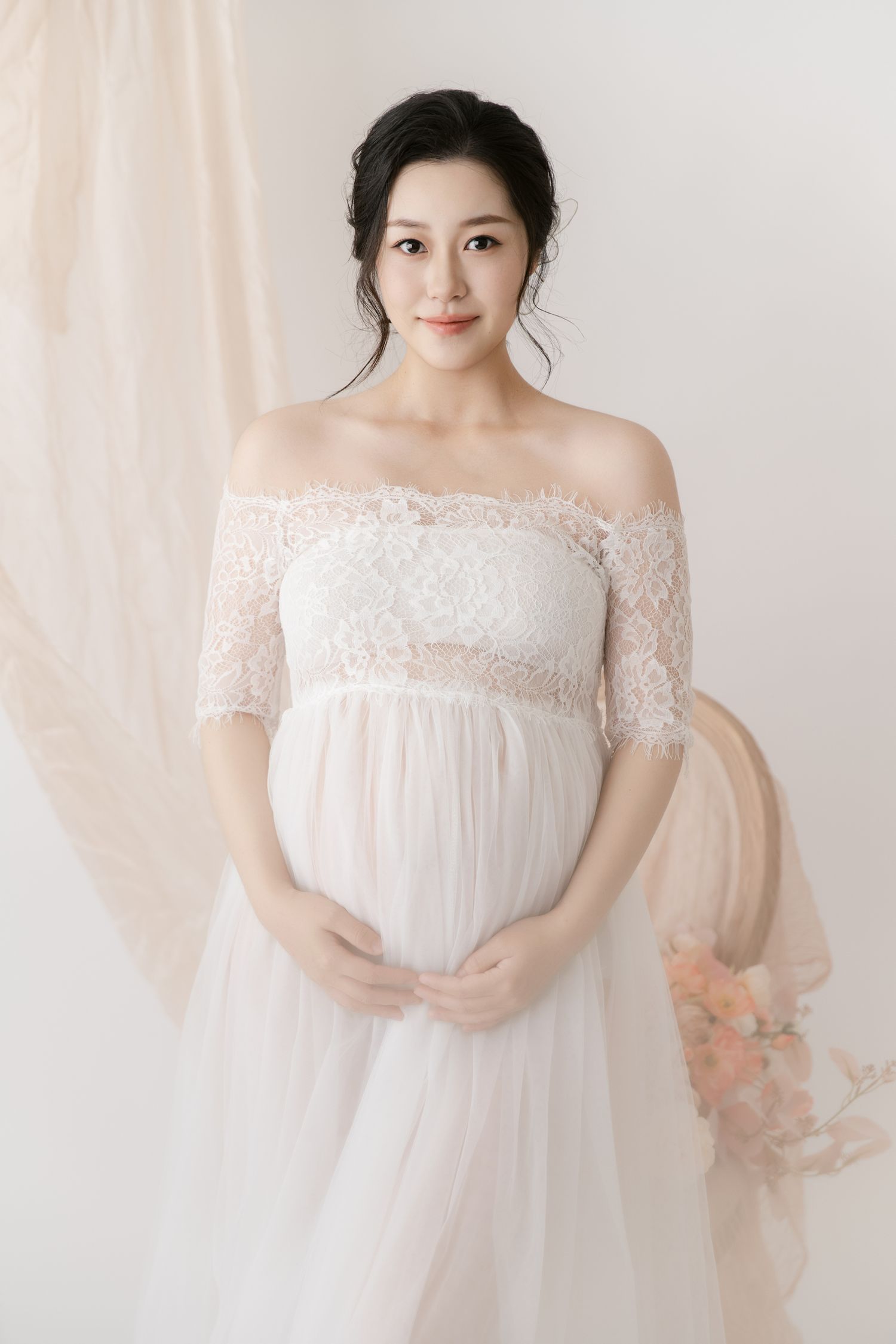 Romantic maternity photos featuring off-shoulder lace dress in delicate white studio setting.