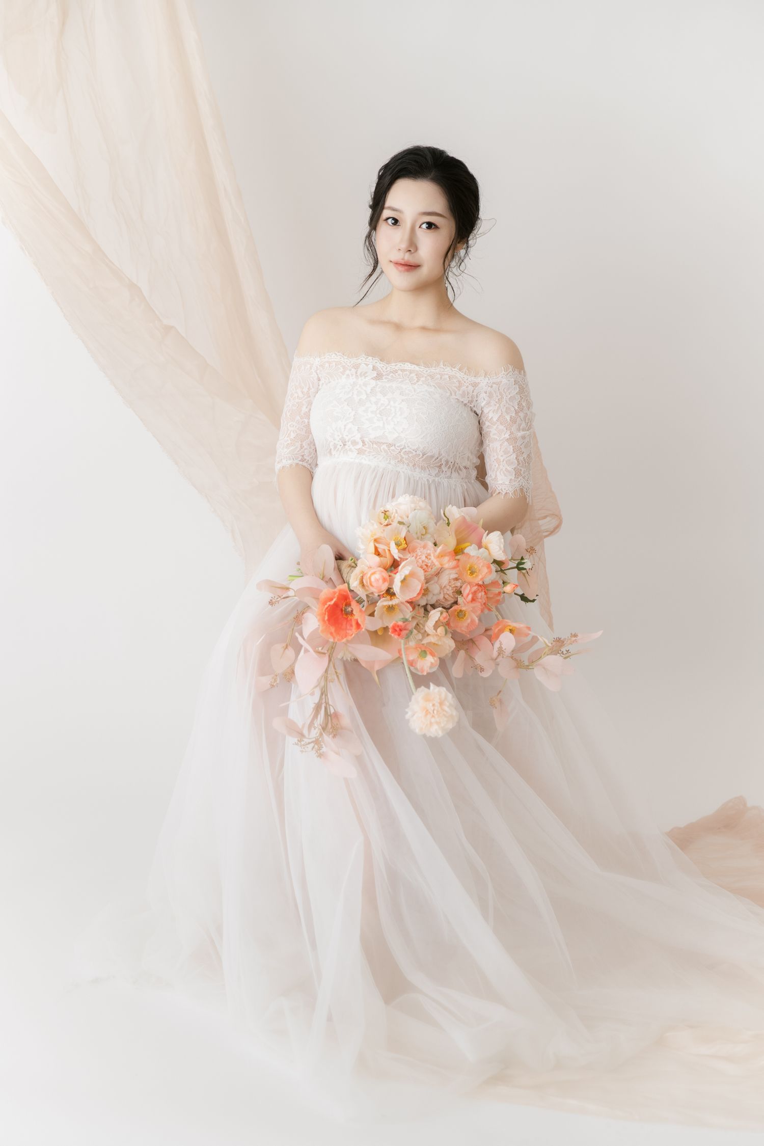 Dreamy maternity portraits with lace dress and coral flower bouquet against light backdrop.