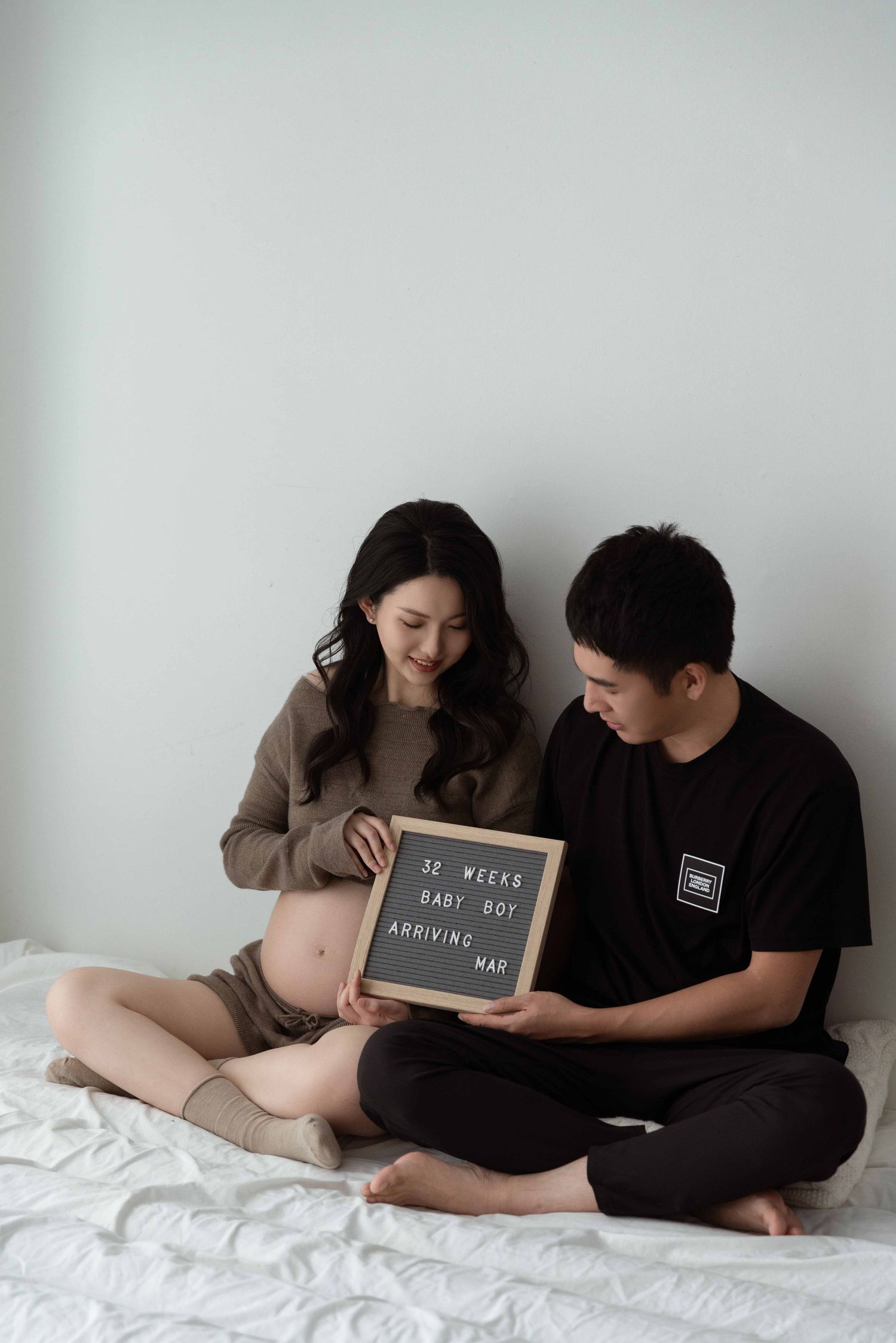 Couple sitting on white bed holding chalkboard signs during maternity photoshoot.